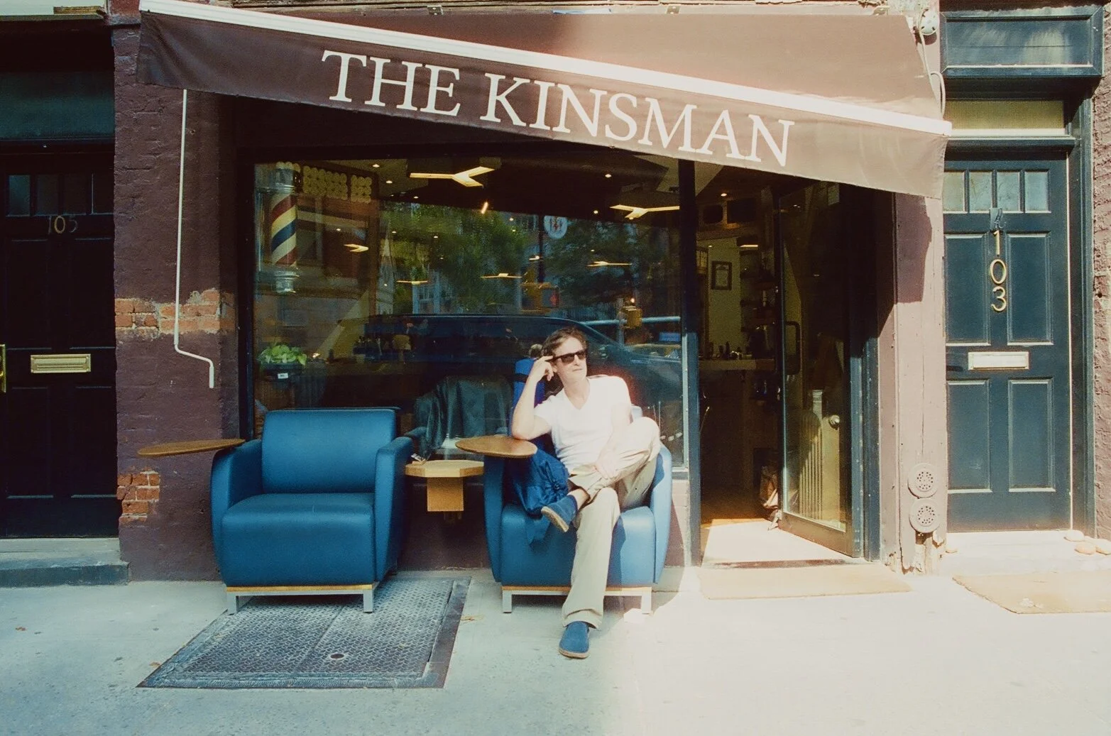 Person sitting outside a cafe named 'The Kinsman', wearing sunglasses, white shirt, beige pants, and blue shoes, with a blue armchair and small wooden tables in front.