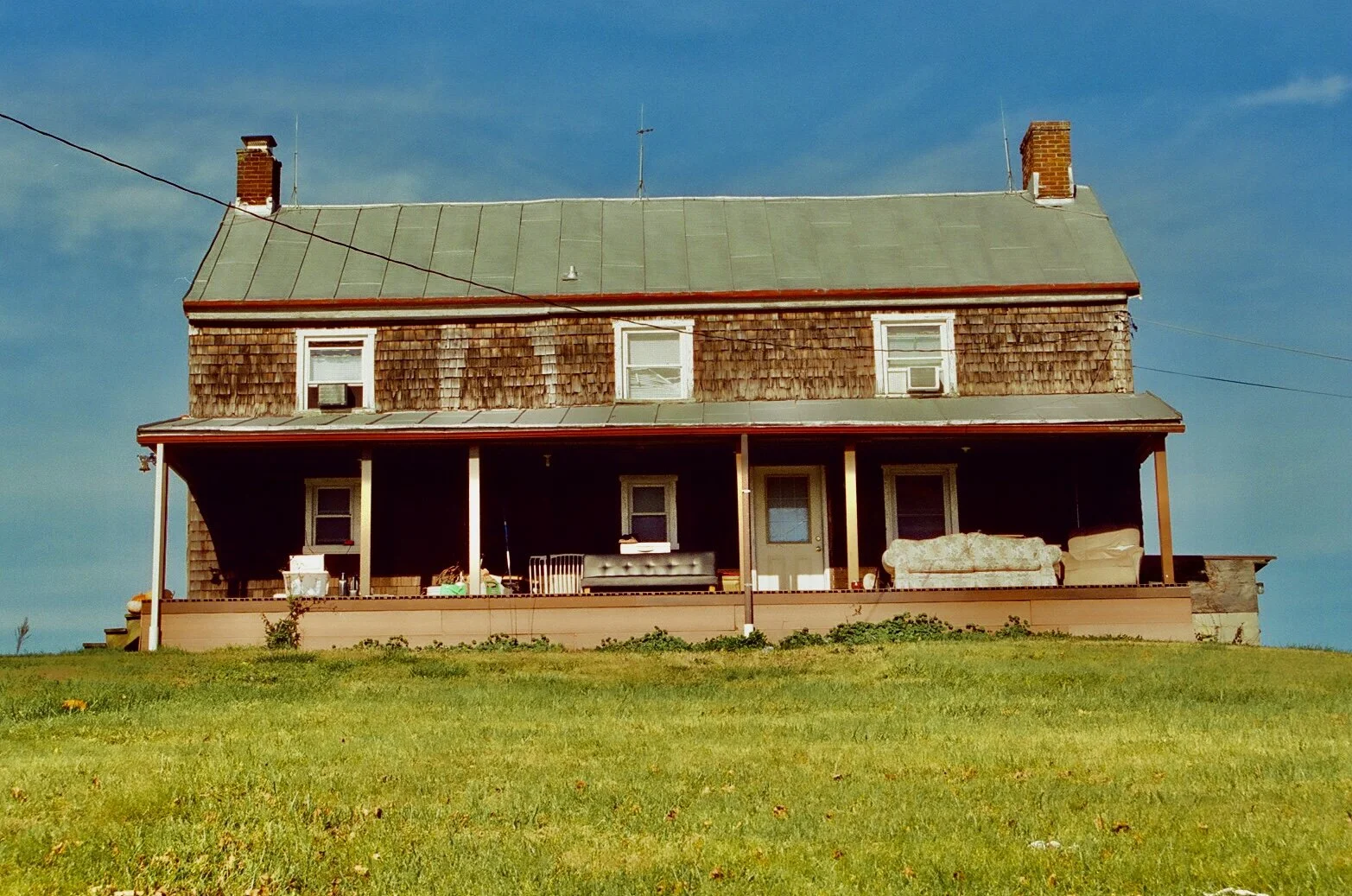 A two-story house with a shingled exterior, greenish roof, and a front porch with furniture, on a grassy hill under a blue sky.