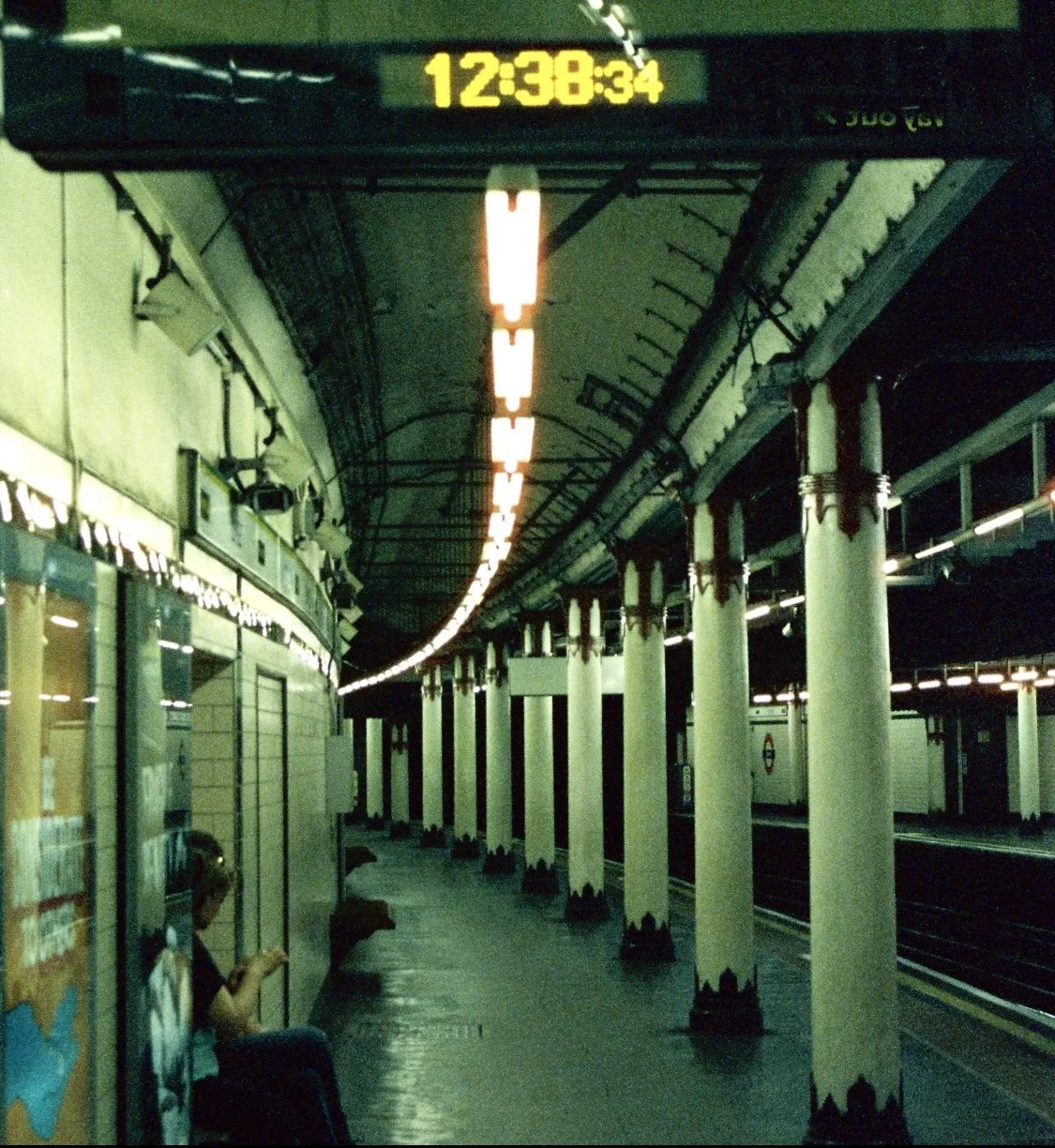 An empty underground subway station with a digital clock displaying 12:38:34, columns with decorative tops, and a person sitting on a bench using a smartphone.