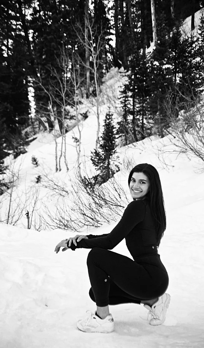 A smiling woman in athletic clothing poses in the snow with a forested hillside in the background.