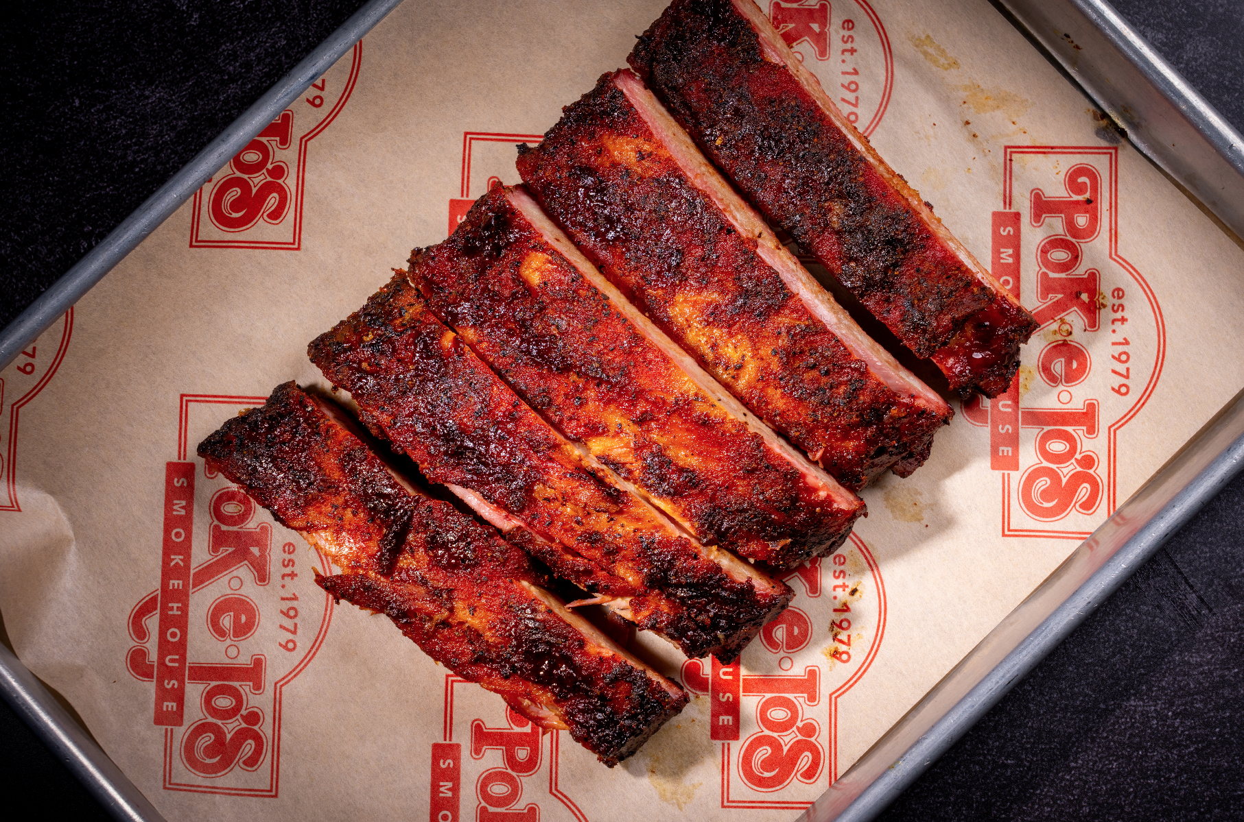 A tray of cooked pork ribs with a smoky, spicy crust, resting on branded paper inside a metal container.
