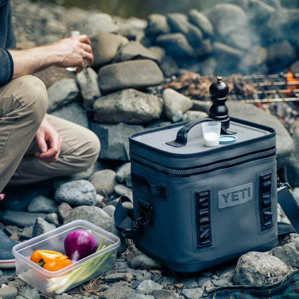 A person camping on rocky ground with a YETI cooler, a small plastic container of vegetables, and a cup of milk on top of the cooler. In the background, there is a stone fire pit and a barbecue grill.