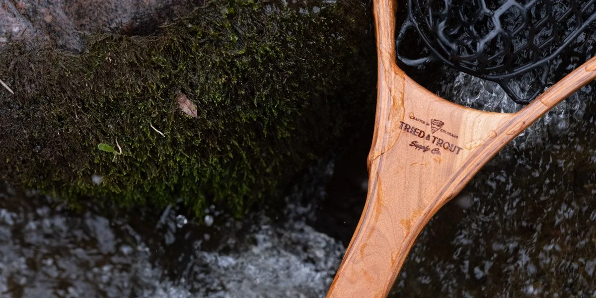 A wooden fishing net with the brand name 'Tried & True Trout' resting in a shallow stream next to a moss-covered rock.