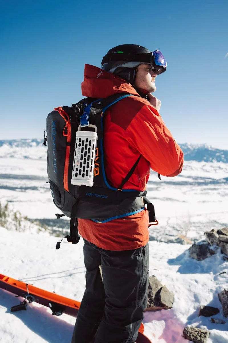 A person in winter gear with a helmet and goggles standing on snow-covered terrain with mountains in the background.