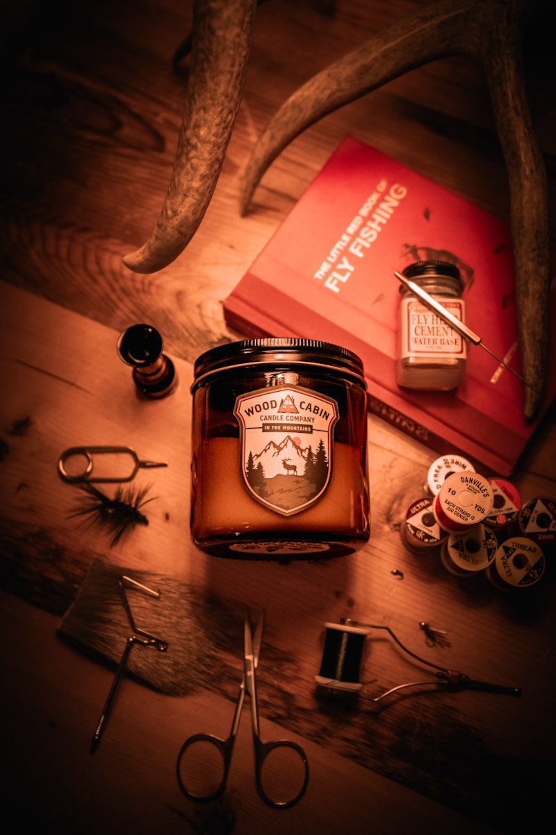 A wooden table with various items for fly fishing, including a candle labeled 'Wood Cabin Candle Company,' a small jar labeled 'Fly Hide Cement,' scissors, a needle, a spool of thread, and containers of fly fishing hooks. An antler and a red book tit