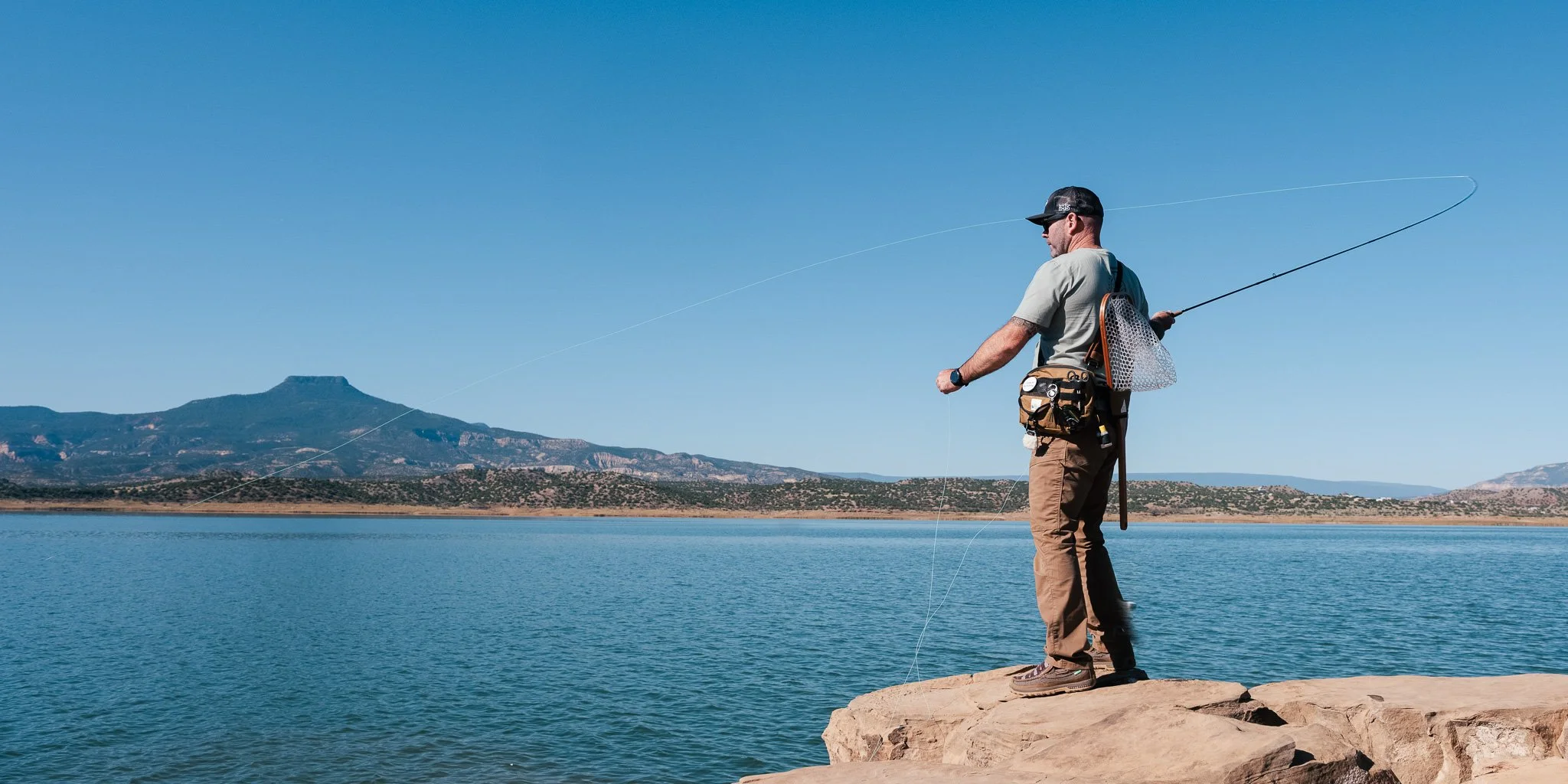 Man fishing on rocky shoreline with mountains and blue sky in background.