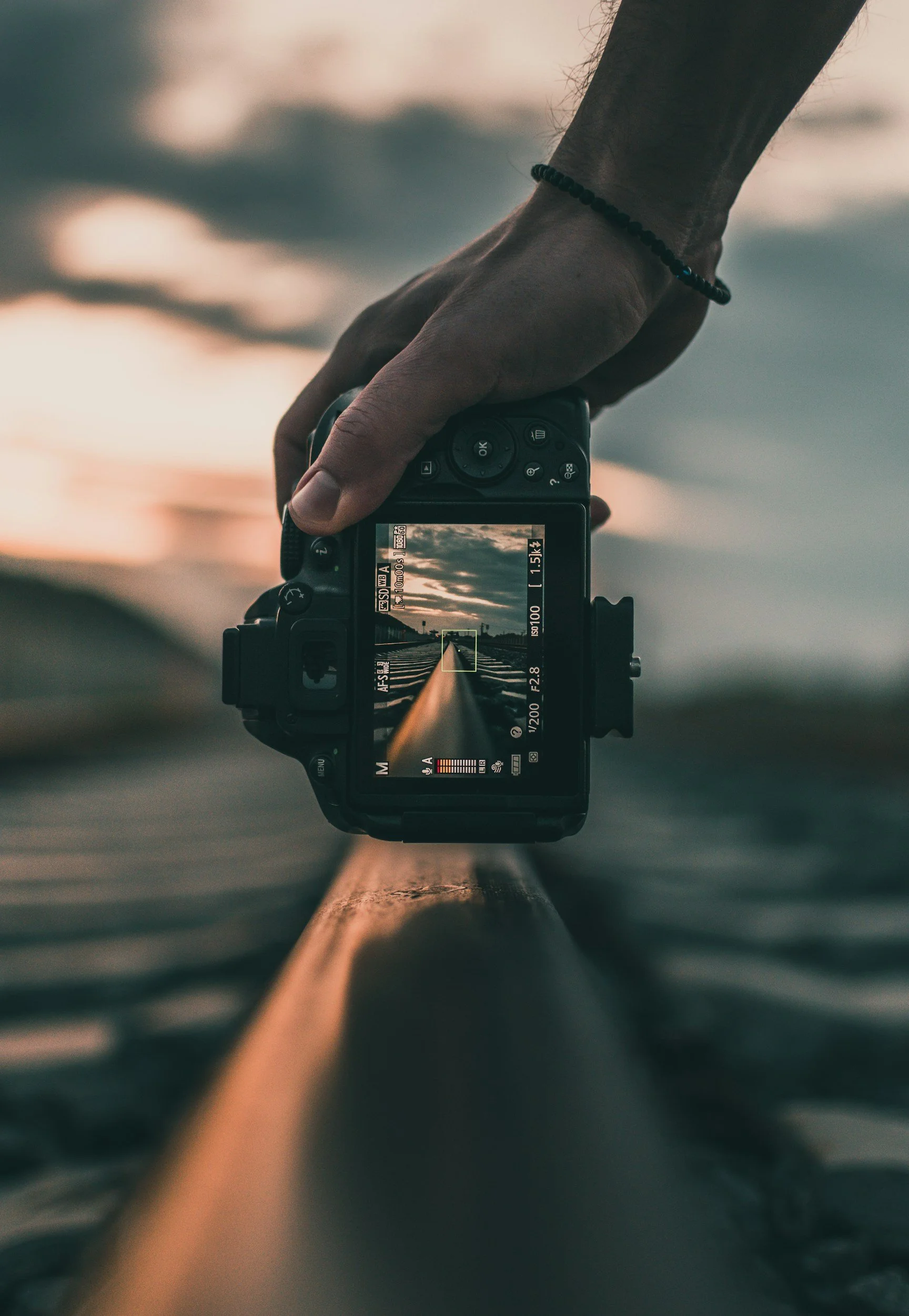 A person holding a DSLR camera over a railway track during sunset, capturing a photo of the tracks extending into the distance.