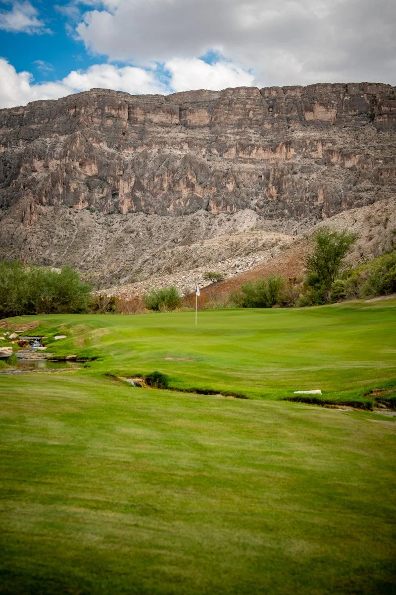 A scenic golf course with a green, a flagstick, trees, and a mountain range in the background under a partly cloudy sky.