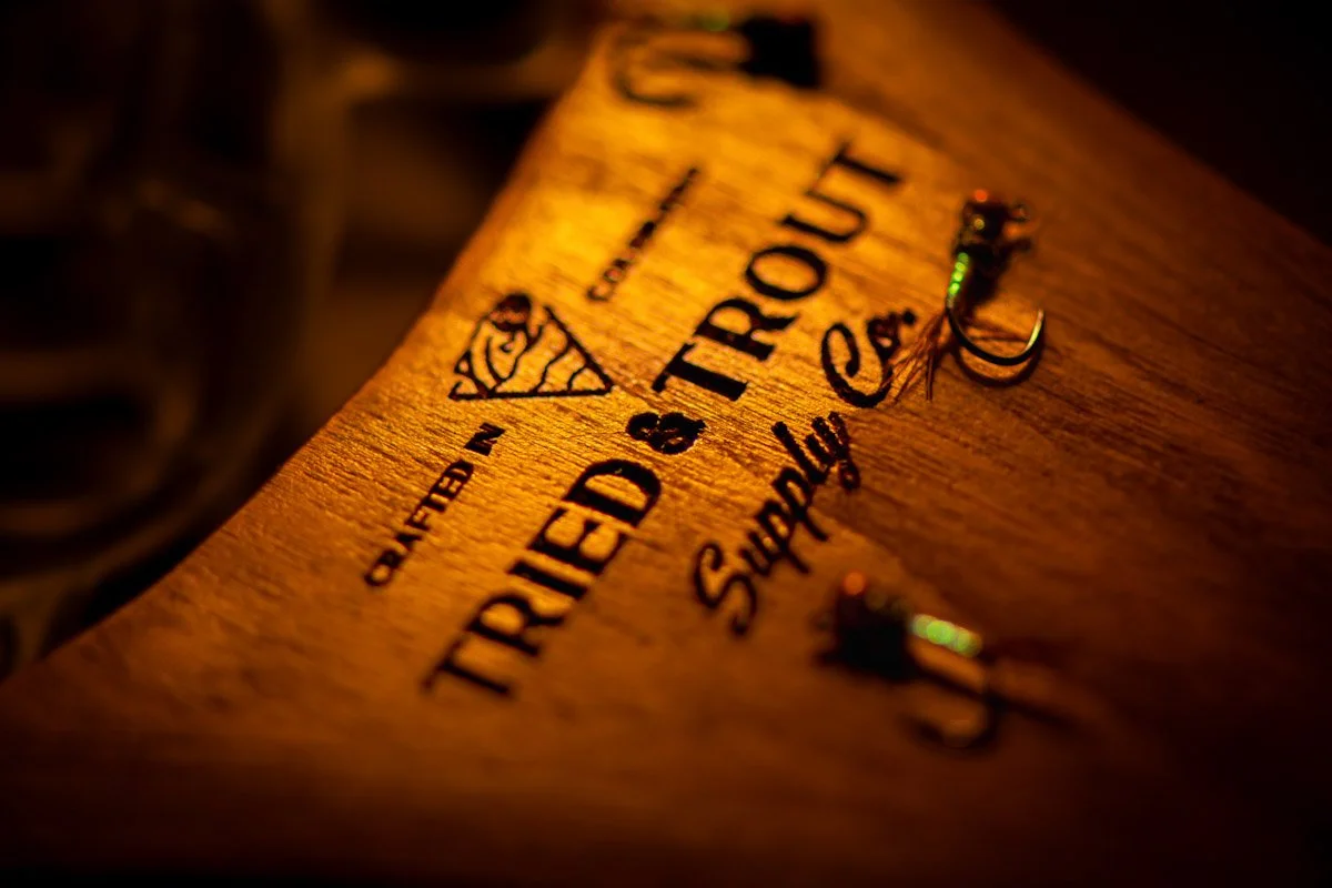 A wooden plaque with black engraved text and a logo, reading 'Taste Your Trip' and 'Supported by'. Two small fishing lures are attached to the plaque with hooks.