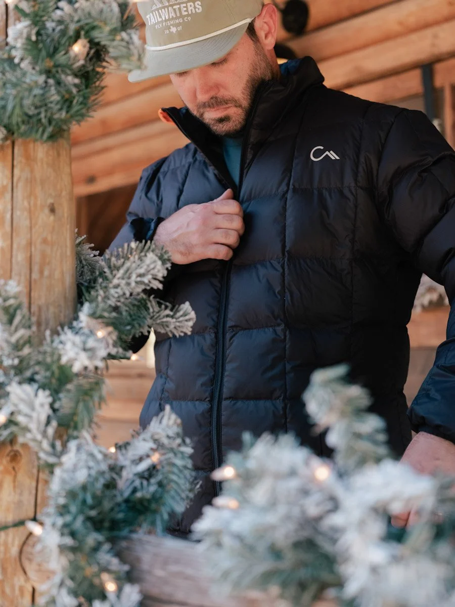A man wearing a gray cap and black puffer jacket, standing among snow-dusted Christmas wreaths and decorations, inside a wooden structure.