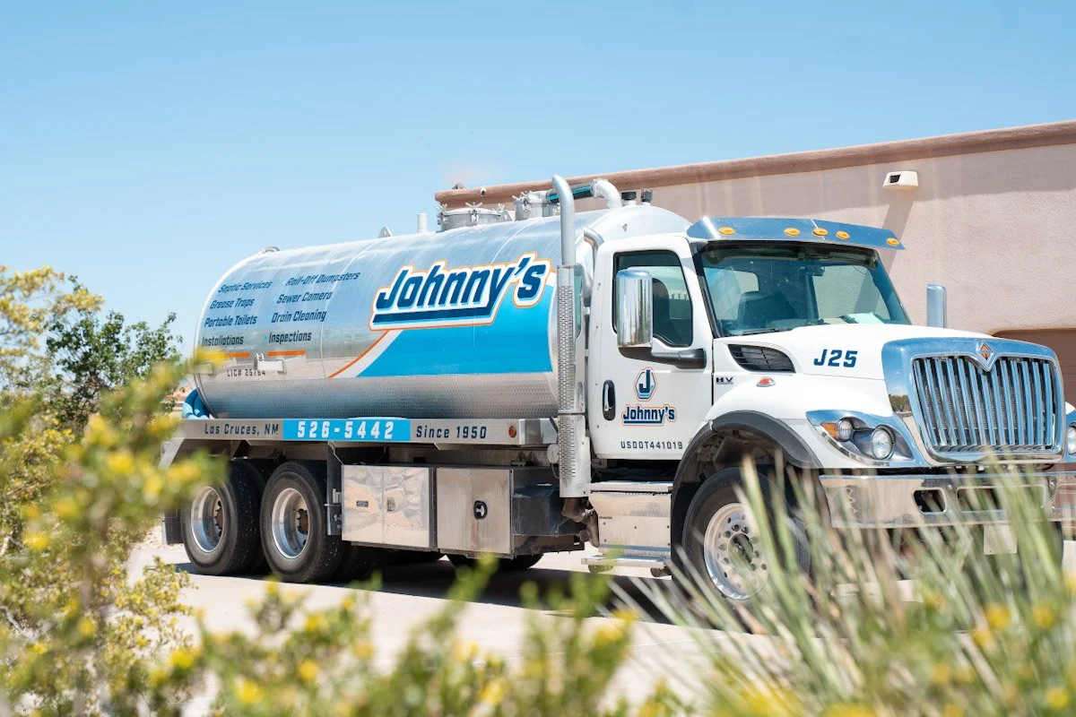 A white Johnny's water truck with blue and orange lettering and a large tank, parked outdoors with plants in the foreground and a beige building in the background