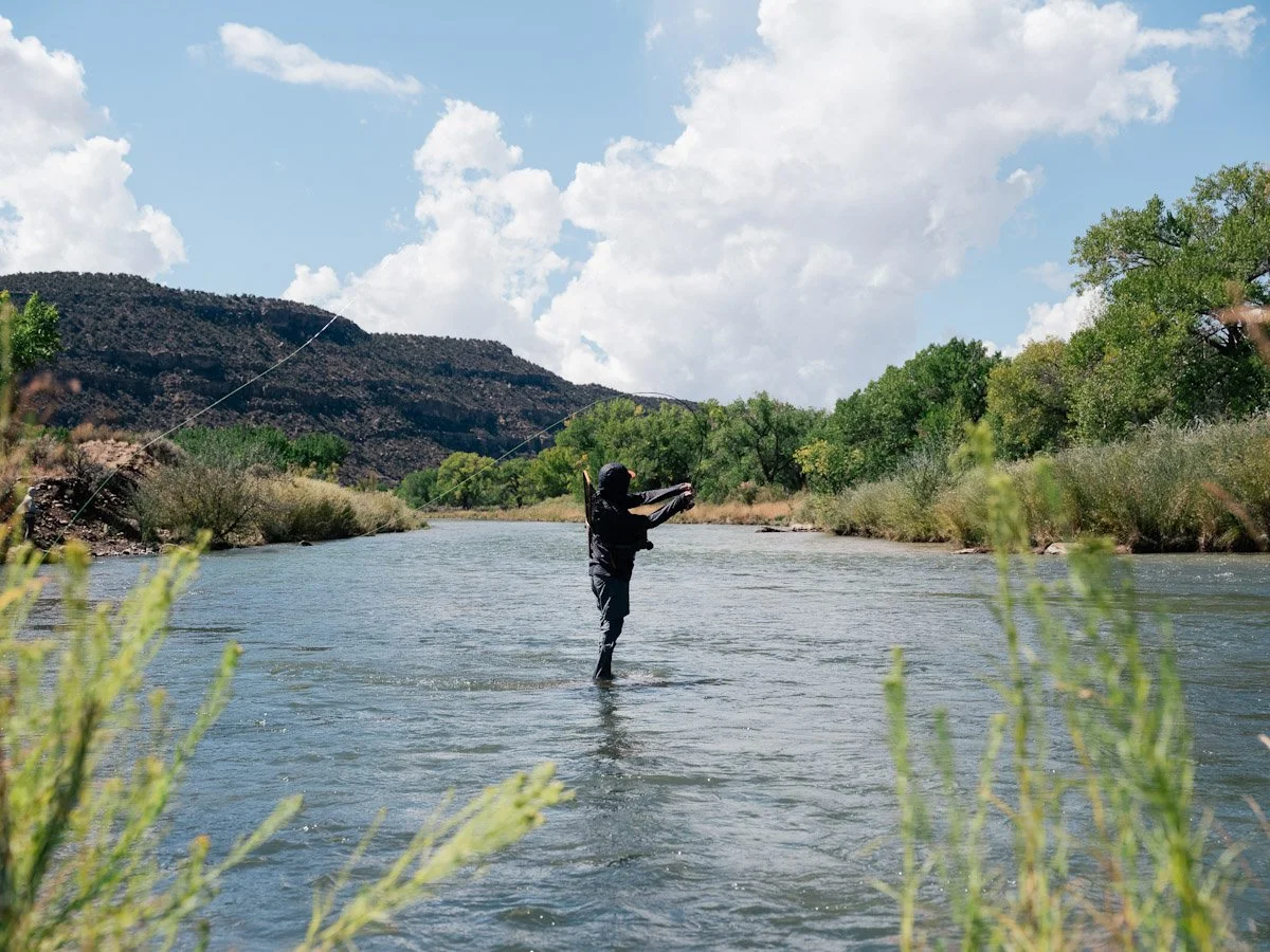Person fly fishing in a river with green trees and mountains in the background under a partly cloudy sky.