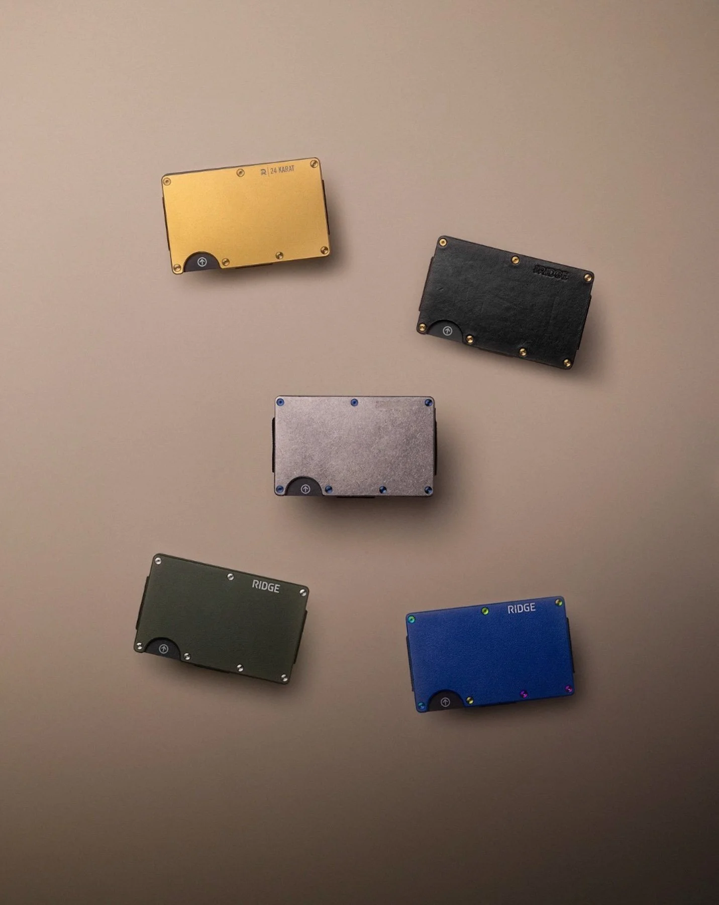 Five small rectangular metal cases with rounded edges in gold, black, silver, green, and blue, arranged on a plain light-colored surface.