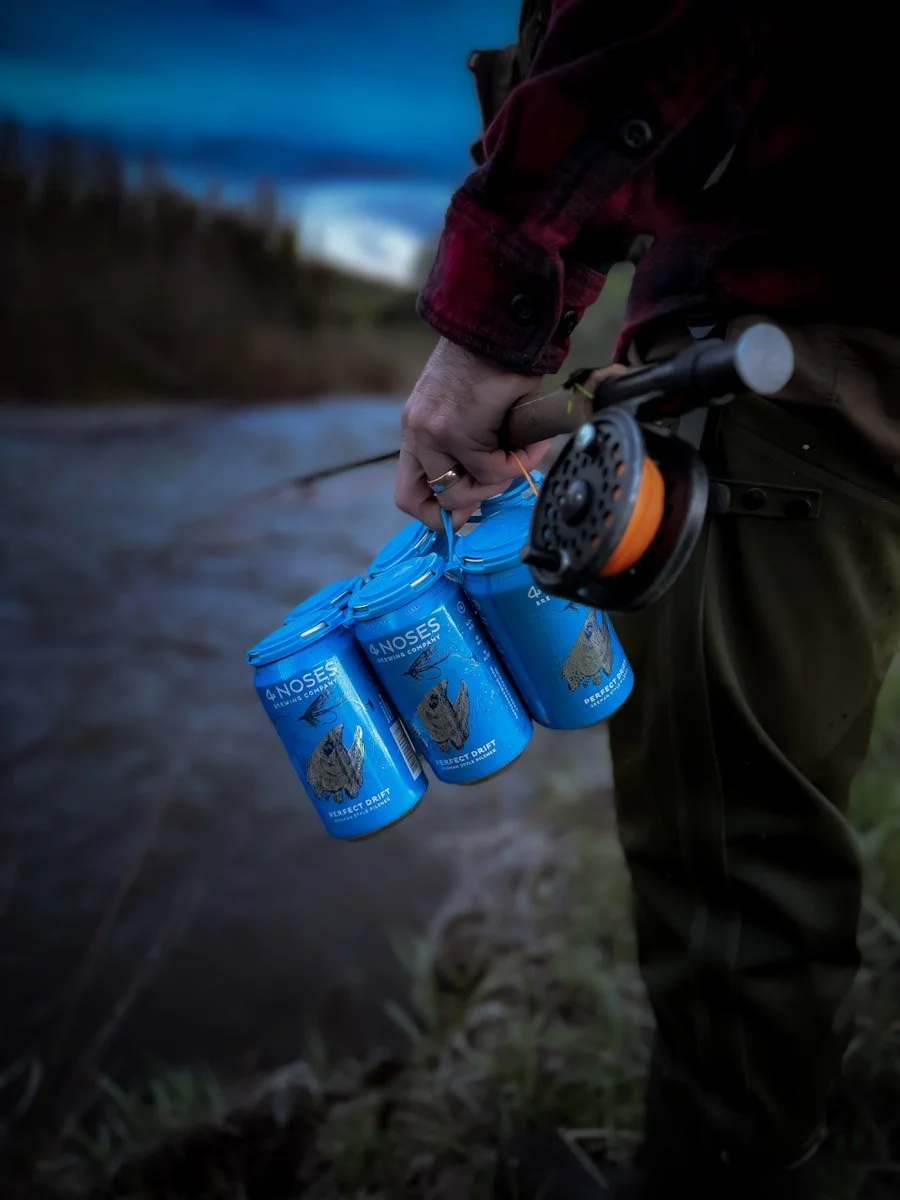 Person holding four blue cans of NOSES sparkling water, with a fishing rod in the other hand, standing outdoors near a river or lake at dusk.