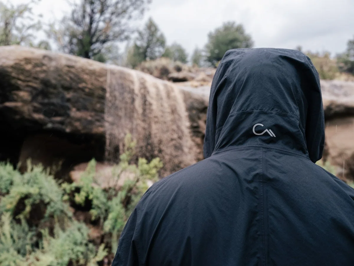 A person wearing a dark jacket with a hood, standing outdoors near a small waterfall or stream, with rocks and sparse vegetation in the background.