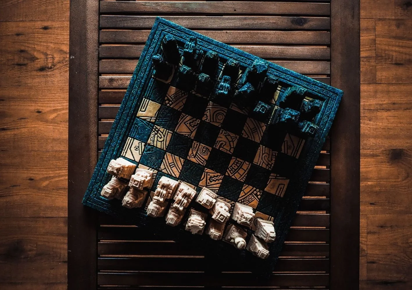 An overhead view of a chessboard with carved stone chess pieces, on a wooden table.