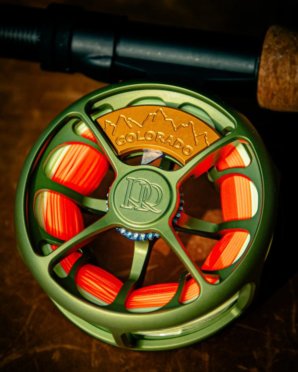 Close-up of a green and orange Colorado-themed roller skate wheel with a mountain logo and the word Colorado, placed on a wooden surface with part of a black object in the background.