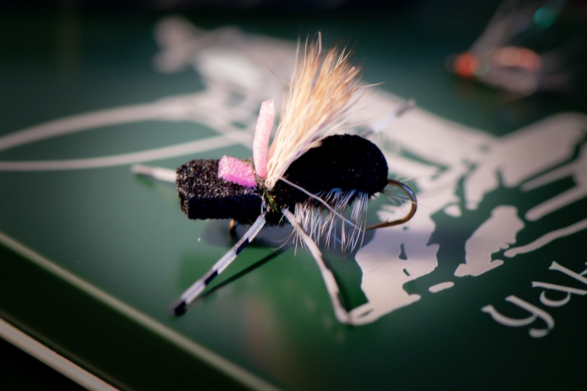 Close-up of a fishing fly with black and pink coloring, attached to a hook, placed on a green surface with a white logo or design.