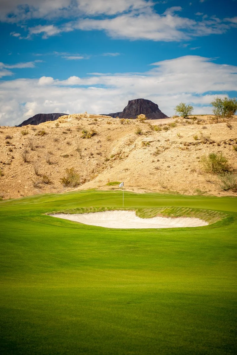 A golf course green with a sand bunker and flag, set against desert hills and rocks under a partly cloudy sky.