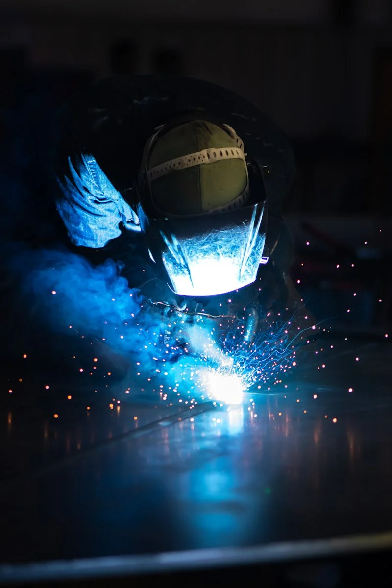 A worker welding, wearing a protective helmet and gloves, creating bright sparks and blue light in a dark environment.