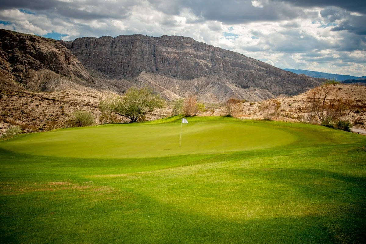 A golf course with a green, nearby trees, and mountain range in the background under a cloudy sky.