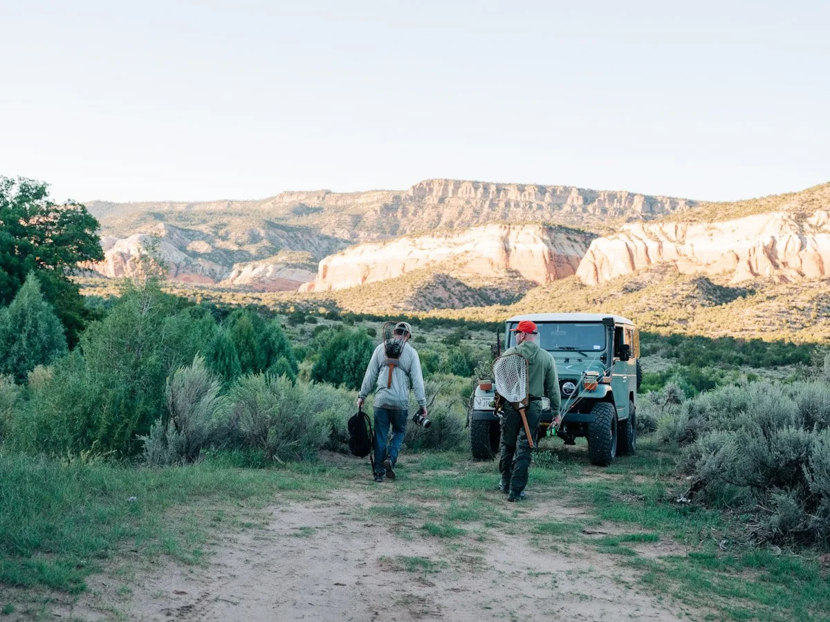 Two people walking on a dirt trail with a Toyota Land Cruiser parked nearby in a scenic desert landscape with mountains in the background.