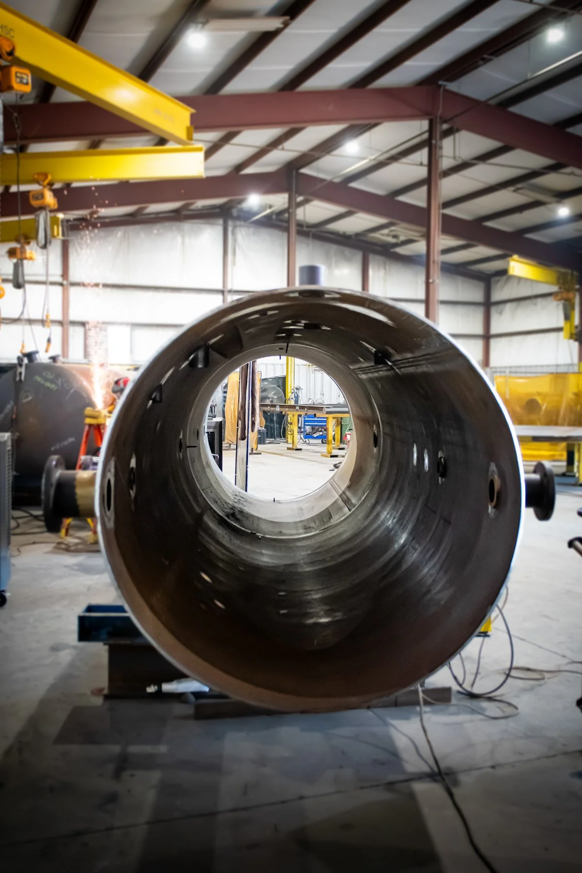 A large metal pipe in an industrial workshop with yellow overhead cranes and tools in the background.