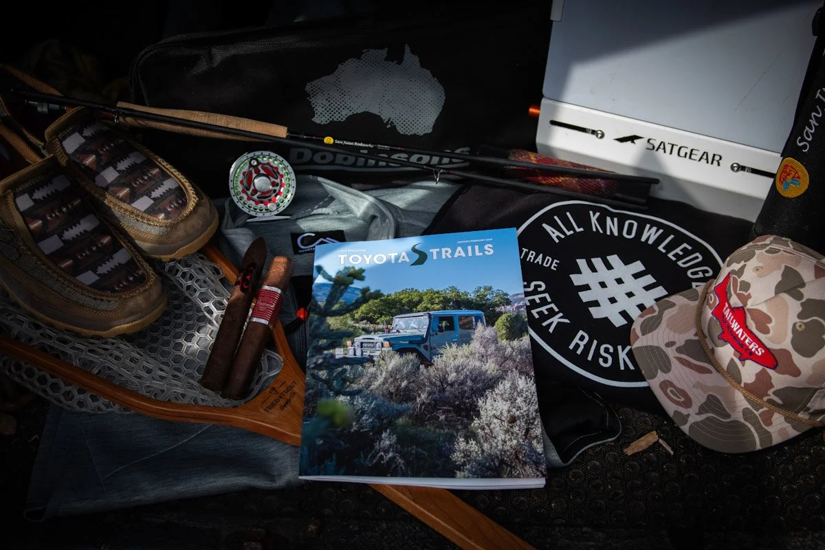 A collection of outdoor and camping gear including a pair of hiking shoes, a cigar, a magazine titled "Toyota Trails" featuring off-road vehicles, a cap with camouflage pattern, a black cloth with white text and symbols, a white box labeled "Satgear," and various other items on a dark surface.