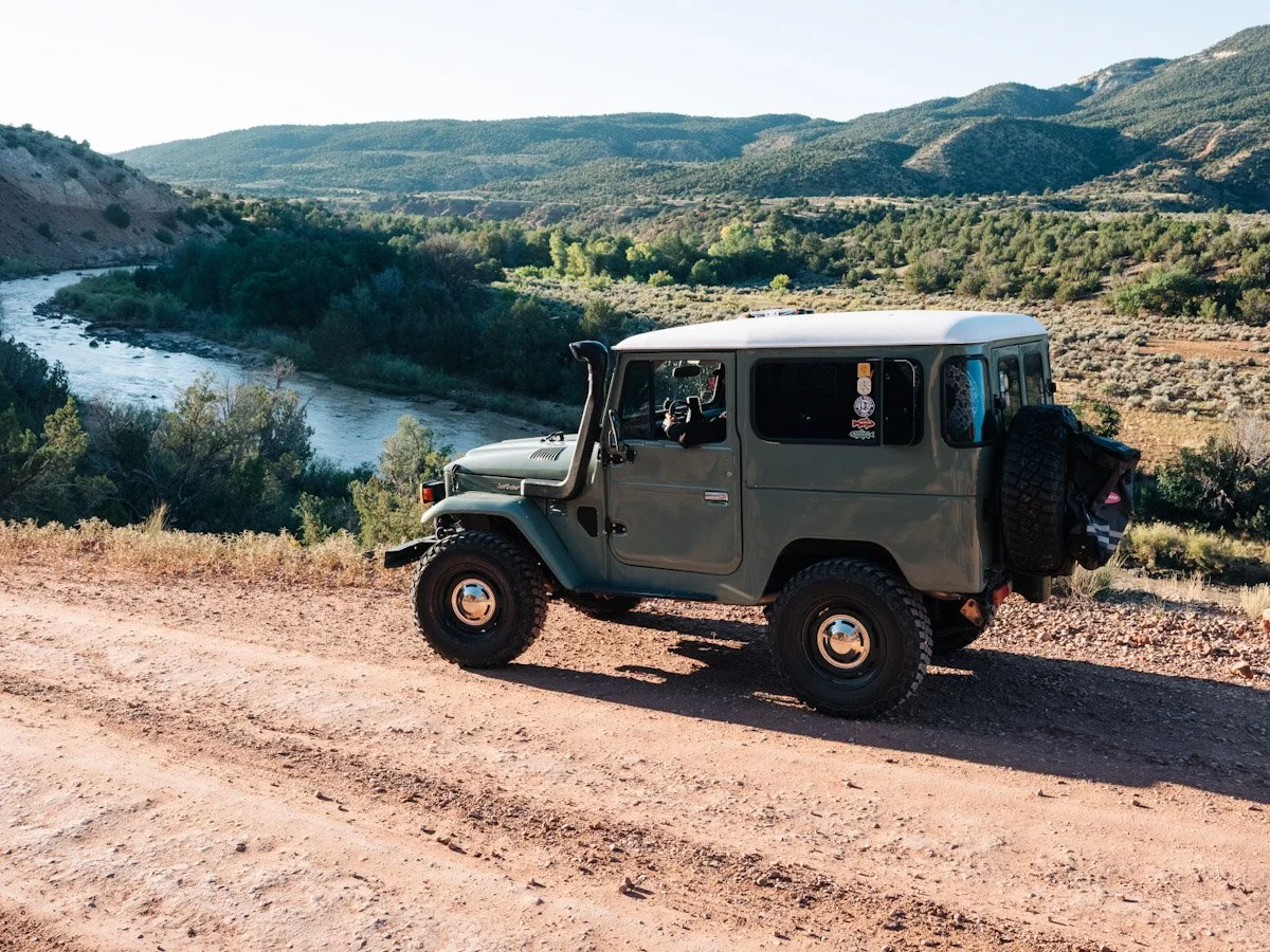 Green off-road vehicle parked on a dirt trail overlooking a river and hills in a desert landscape.