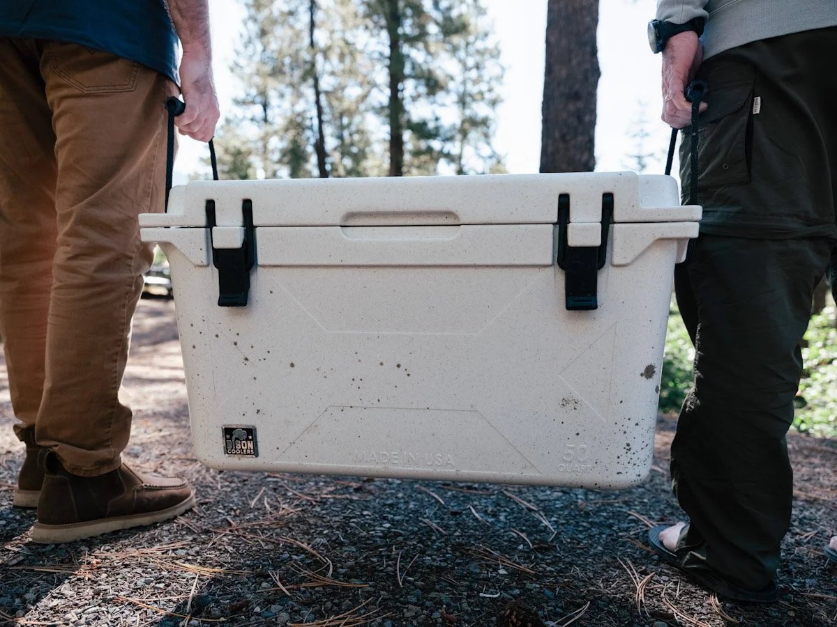 Two people are carrying a large white cooler with black handles through a wooded outdoor area.