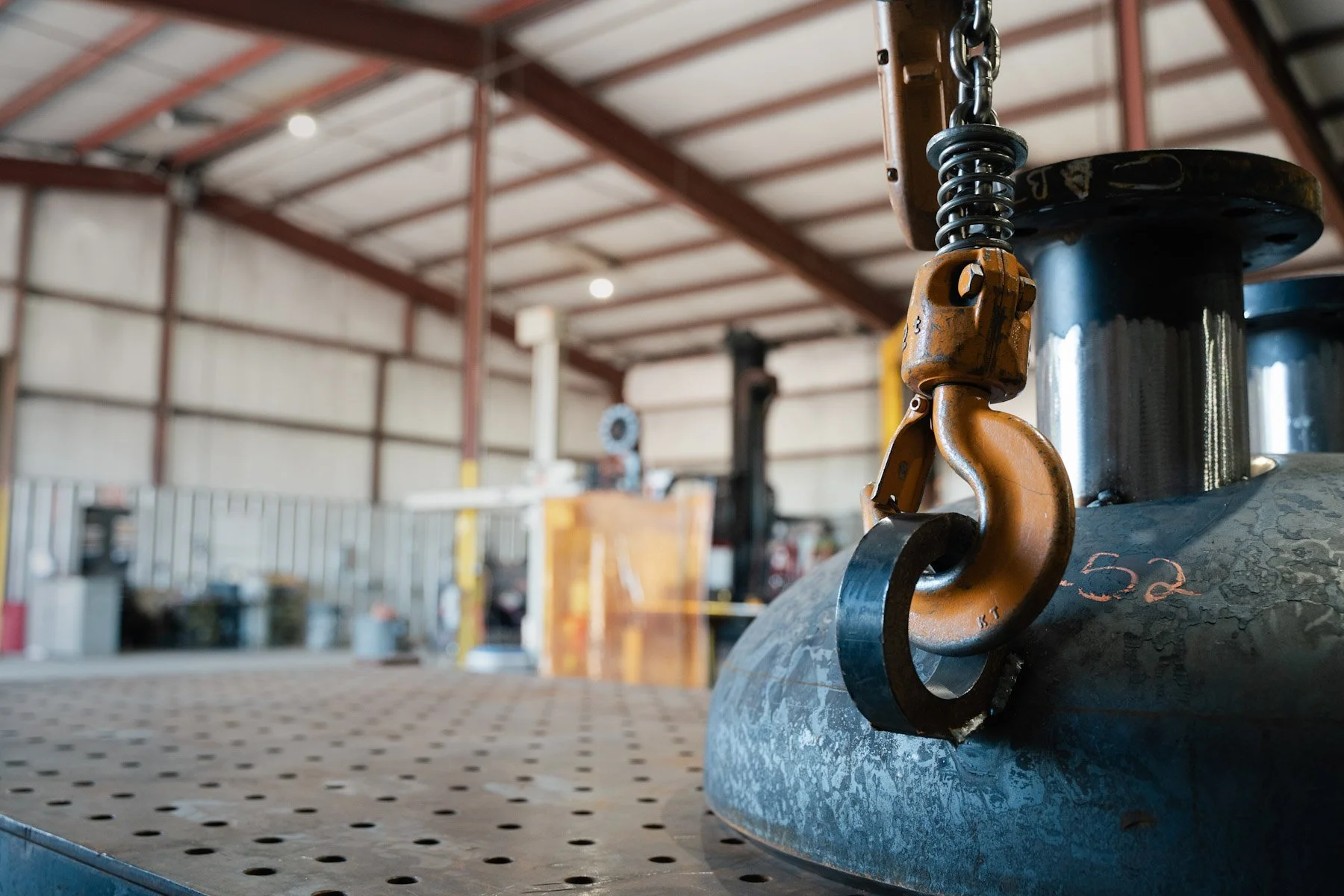 A close-up of a metal hook and chain attached to a cylindrical black tank in a workshop or industrial setting.