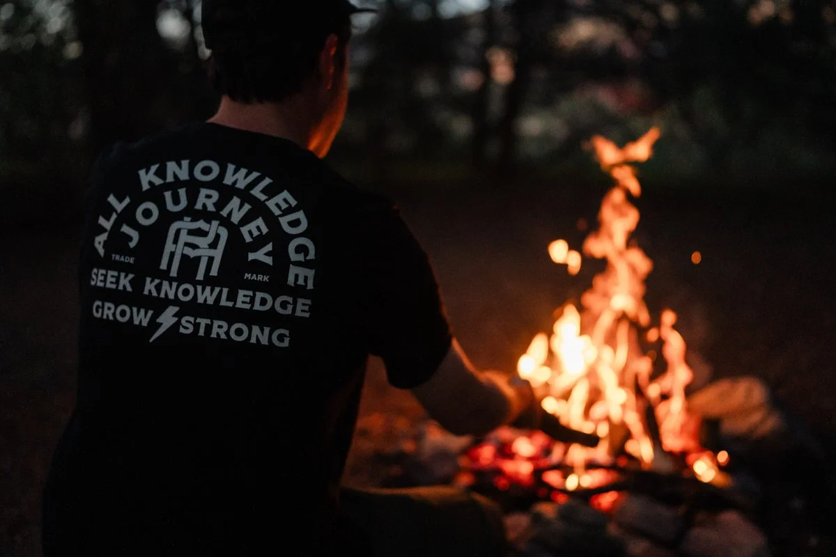 Person cooking around a campfire at night, wearing a black T-shirt with white text.