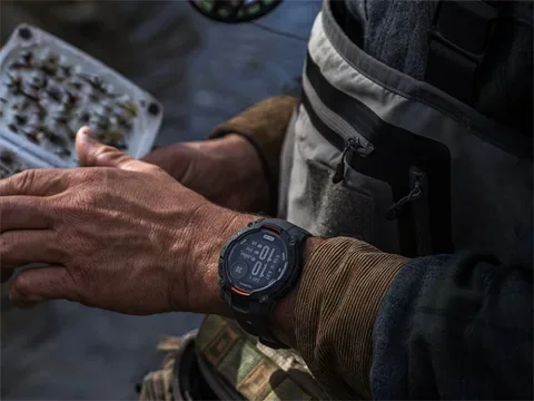 Close-up of a person's left wrist wearing a digital watch, with a weathered hand, next to another person's leg in outdoor clothing and equipment.