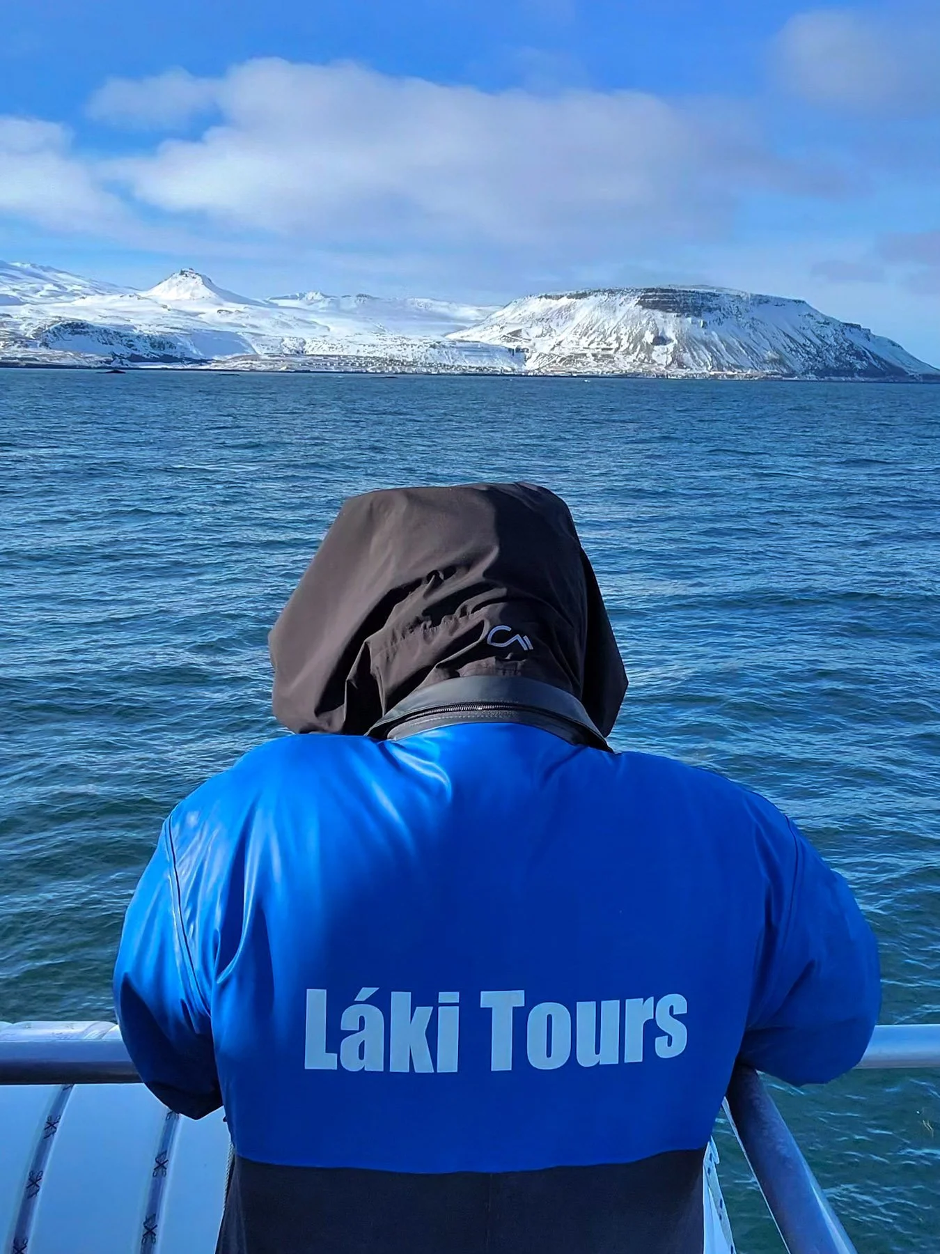Person in a blue jacket labeled 'Láki Tours' standing on a boat, facing away, looking at snowy mountains across the water.