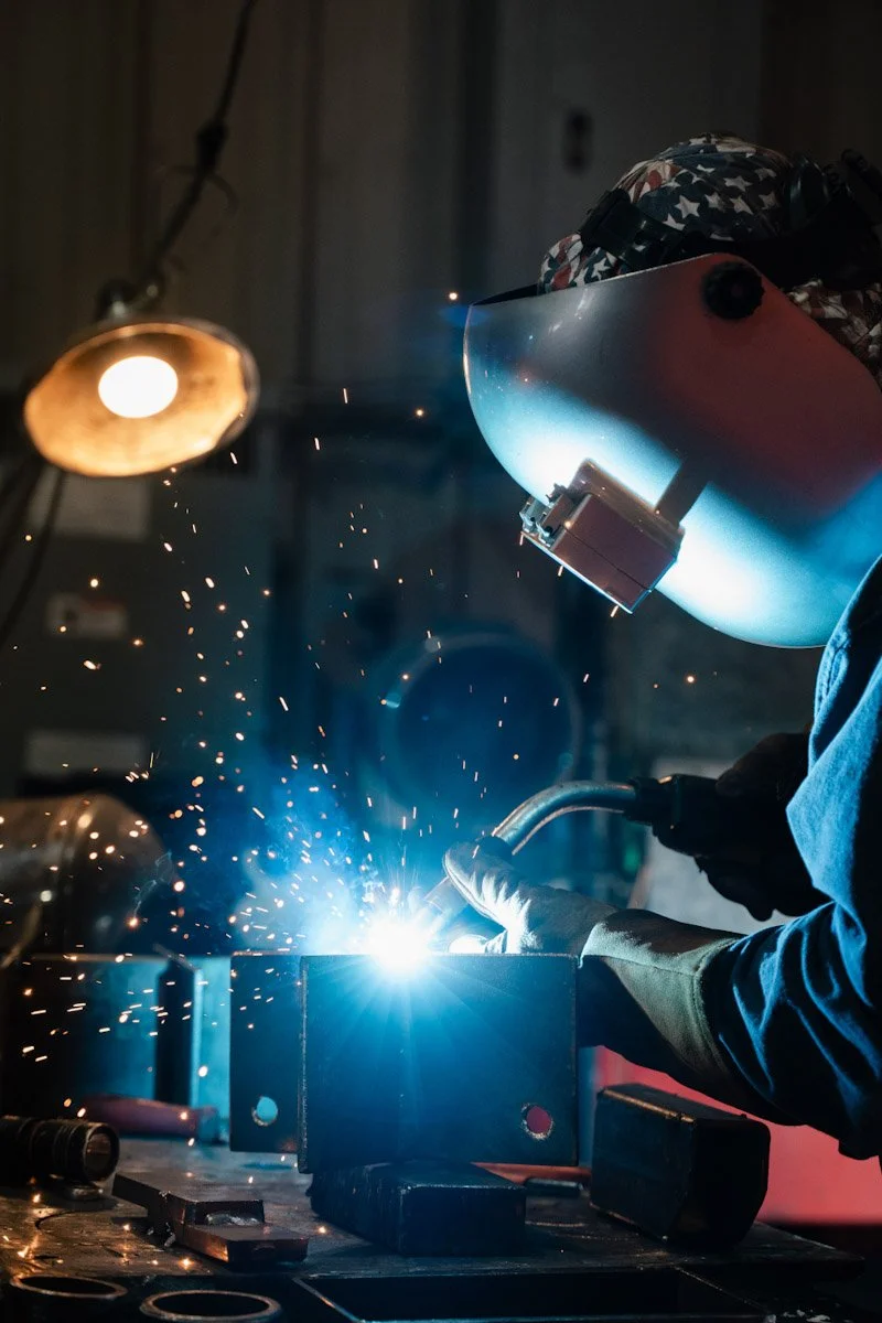 A person welding metal in a workshop, wearing a protective helmet, gloves, and blue clothing, with sparks flying from the welding process.