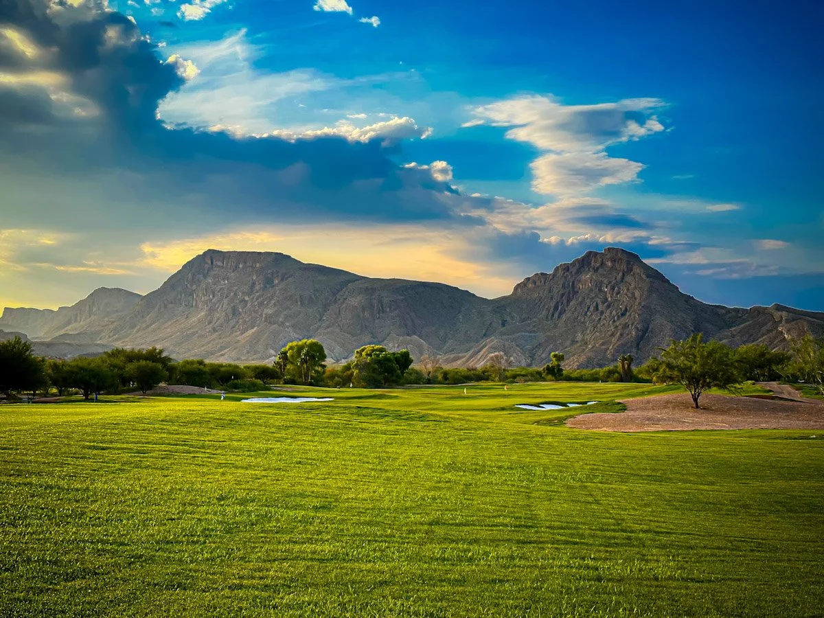 A golf course with lush green grass, sand bunkers, and small water hazards, set against a backdrop of mountains and a partly cloudy sky during sunset.