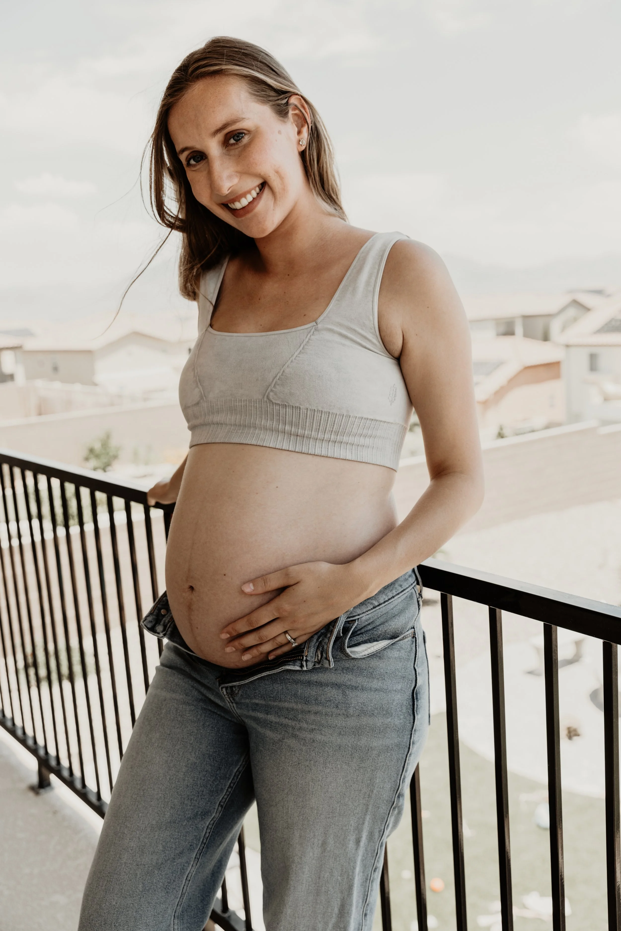 A pregnant woman standing on a balcony, smiling at the camera, wearing a beige crop top and blue jeans, with a residential neighborhood in the background.