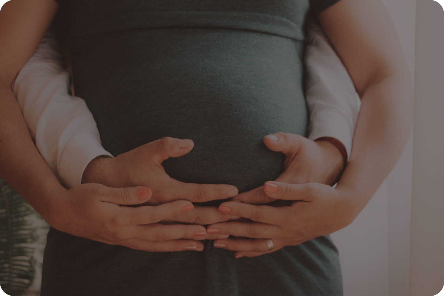 Multiple hands, including that of a father and a mother, gently rest on a pregnant belly, forming a heart shape with their fingers.