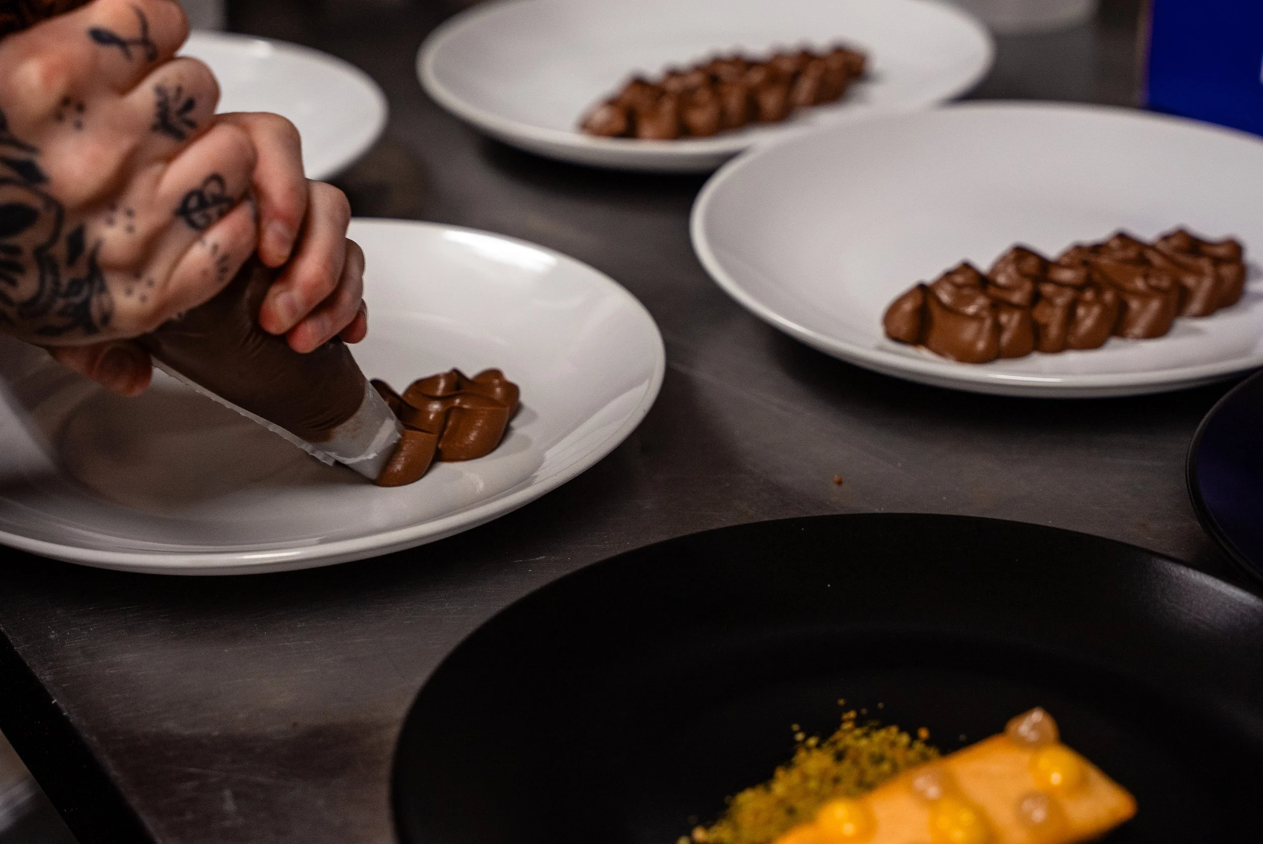 Close-up of tattooed hands squeezing chocolate onto white plates, where similar chocolate confections are arranged.