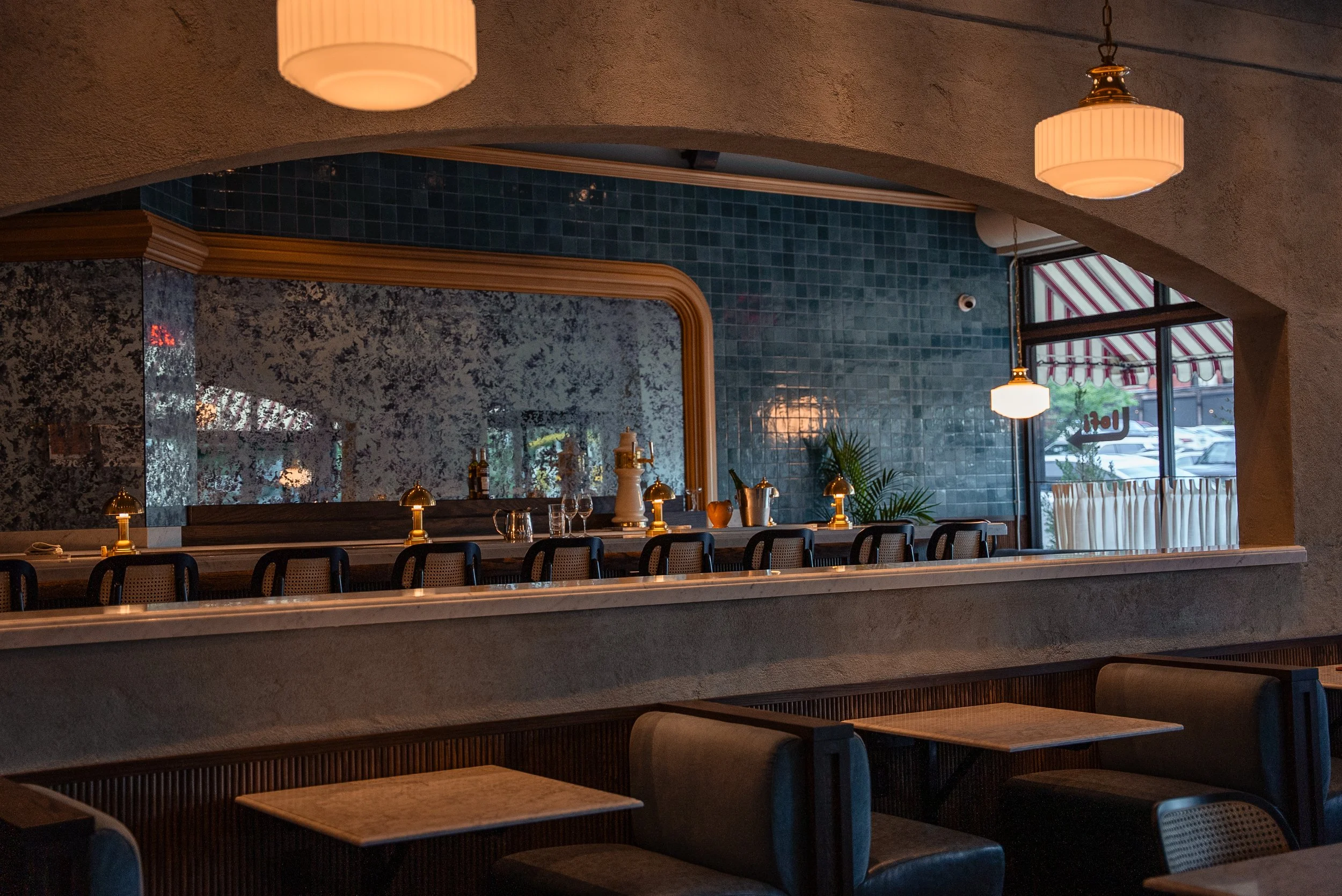 Interior of a restaurant with a bar, black chairs, marble countertops, and hanging pendant lights near large windows with striped awnings.