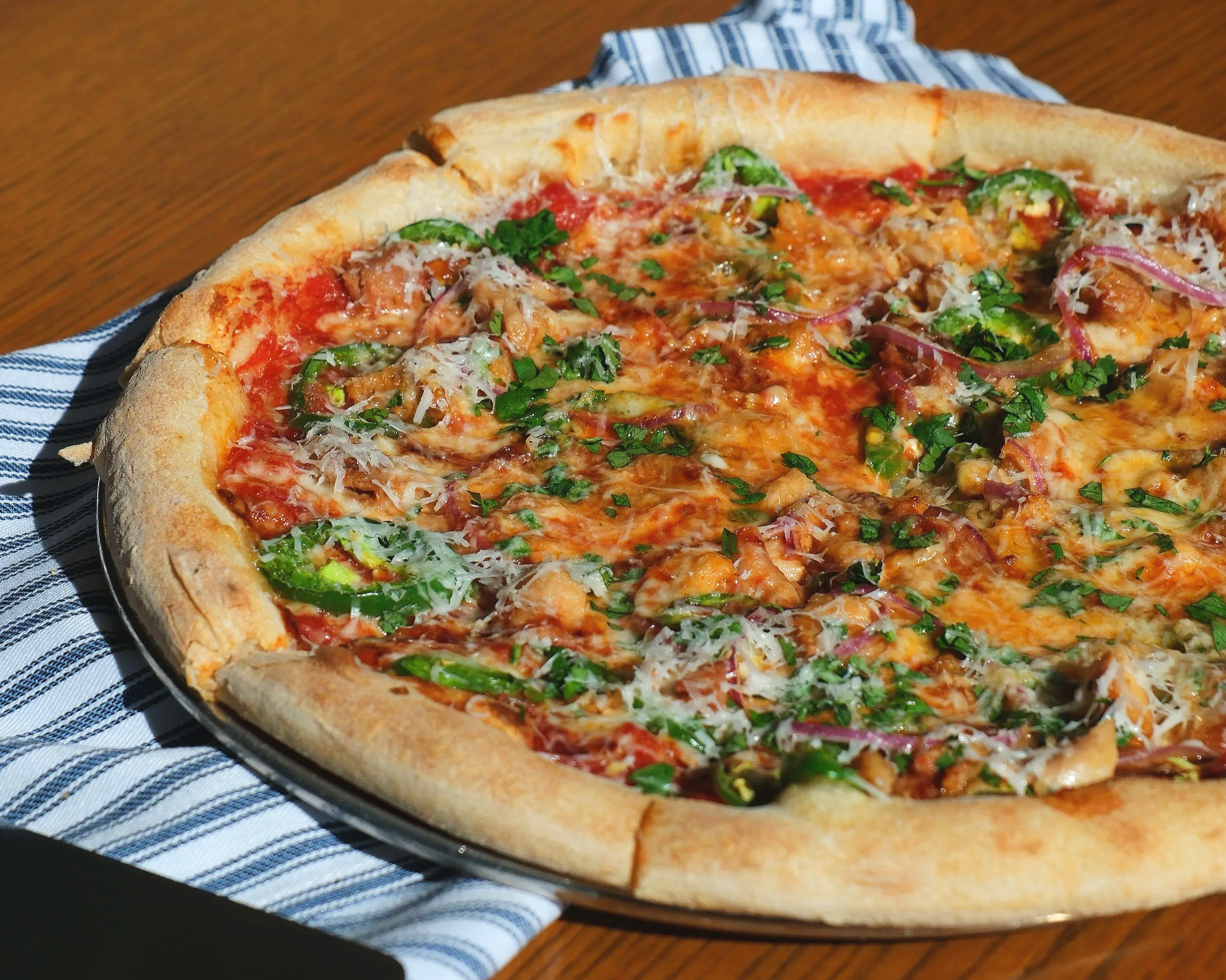 A freshly baked pizza with tomato sauce, cheese, green peppers, red onions, chopped herbs, and shredded cheese on a black plate, placed on a wooden table with a blue striped cloth.