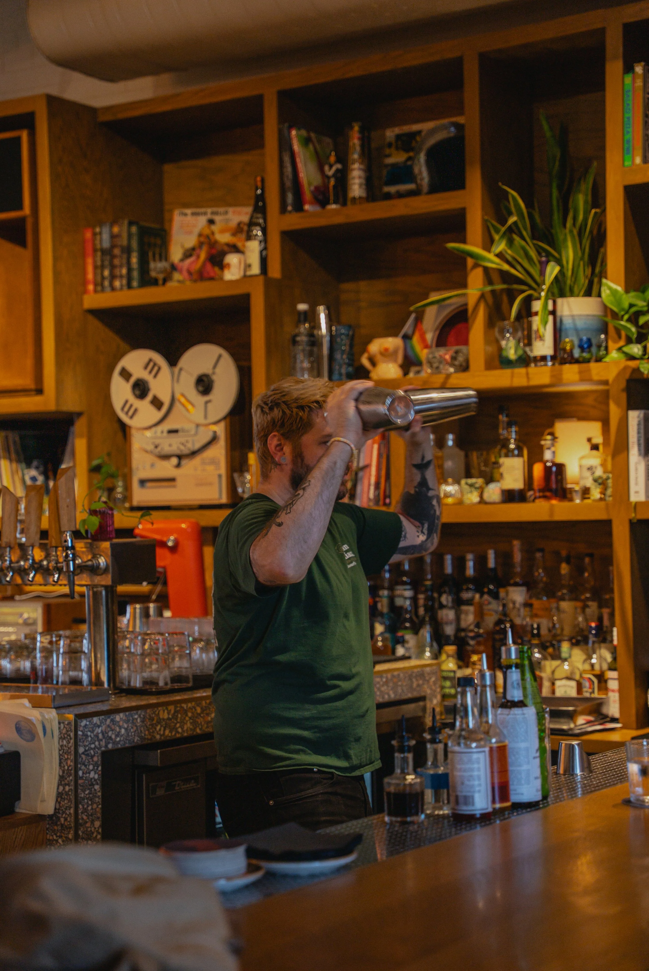 A bartender with tattoos wearing a green shirt is looking through a cocktail shaker behind a bar, with shelves of alcohol, books, and plants in the background.