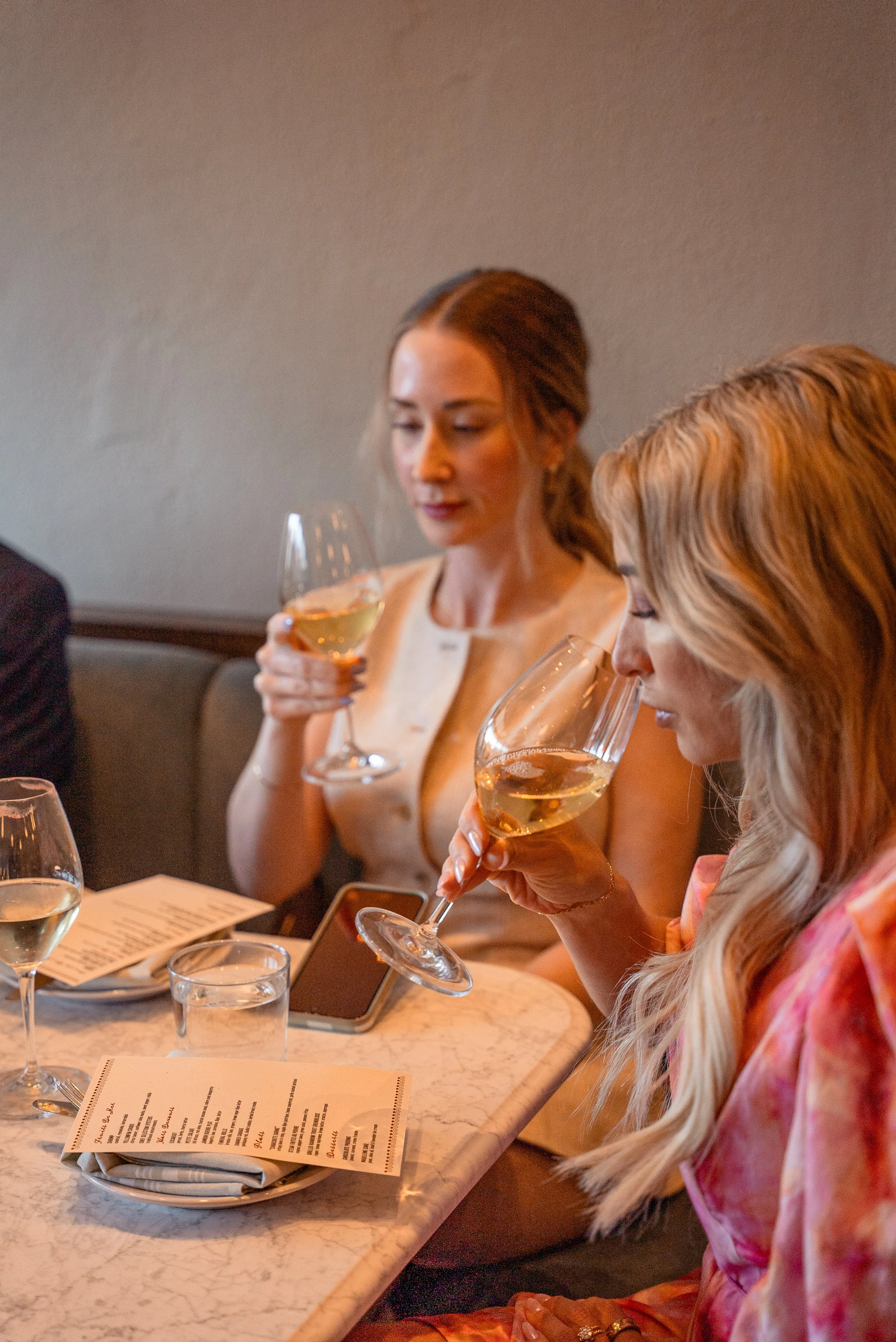 Two women sitting at a table, each holding a glass of white wine in a restaurant or café, with menus and a smartphone on the table.