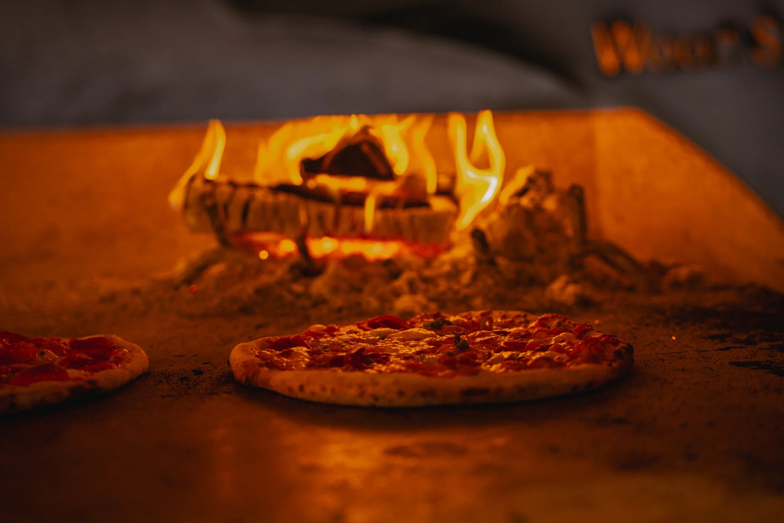 Pizzas baking in a wood-fired oven, with flames visible in the background.