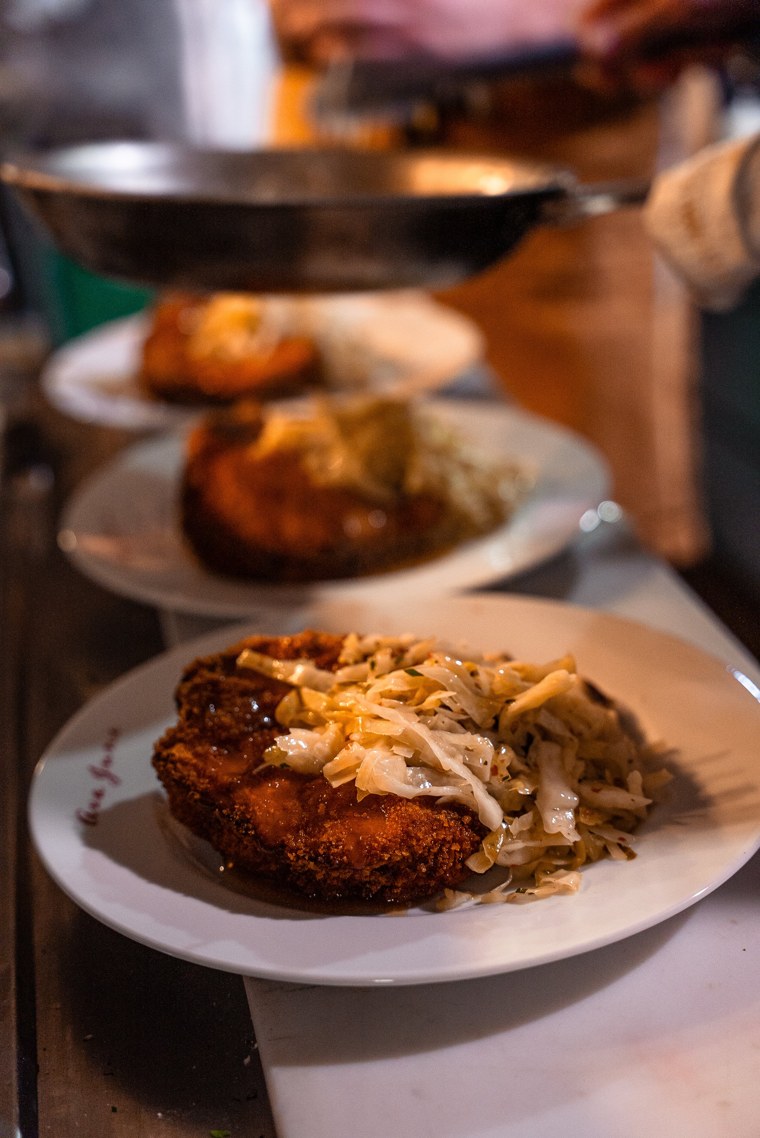 A plate with a breaded fried cutlet topped with coleslaw, set on a table with other plates of food in the background.
