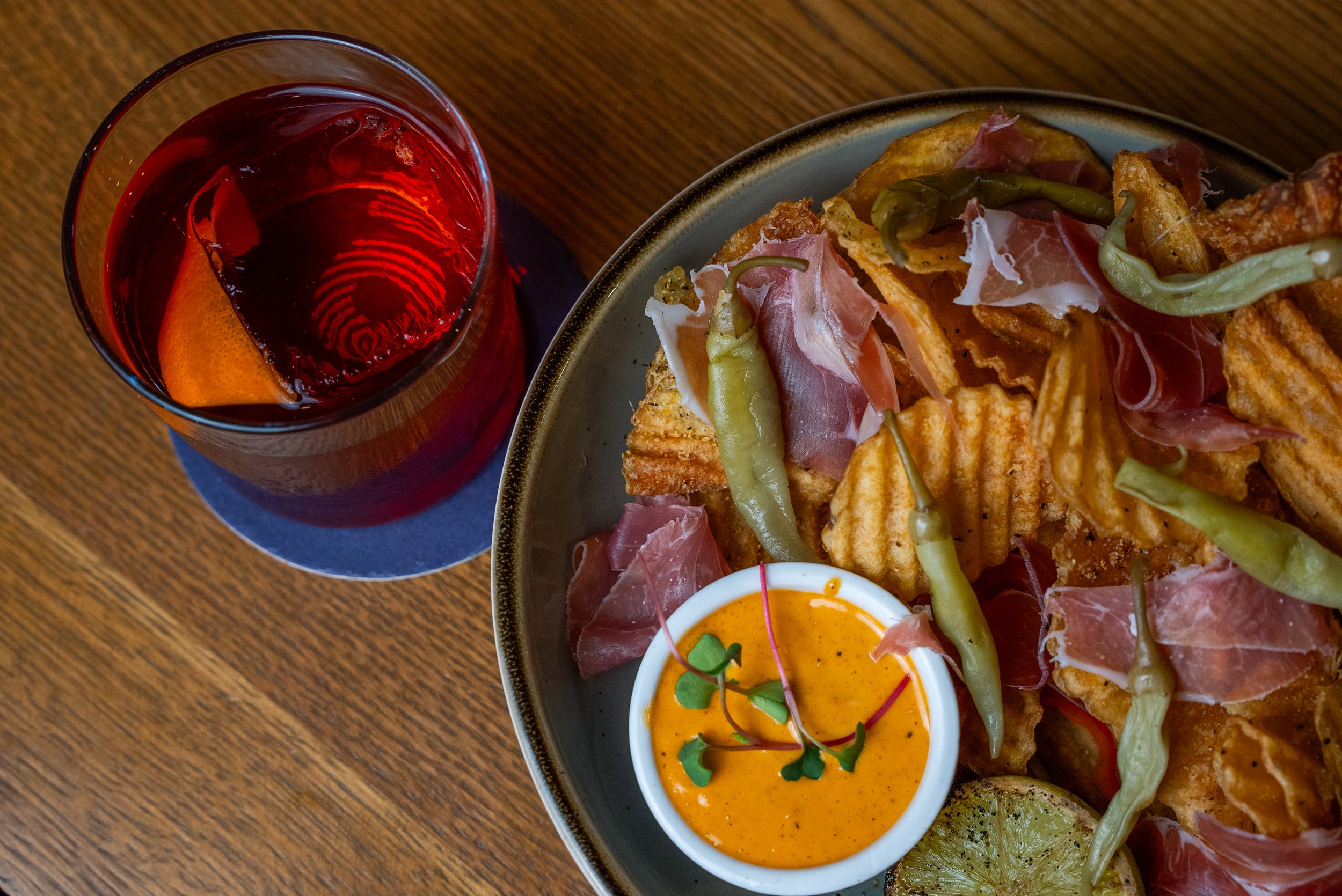 A glass of red drink with ice and an orange wedge, a bowl of waffle fries topped with prosciutto and green chili peppers, a small cup of orange dipping sauce garnished with microgreens, and a slice of lime on a wooden table.