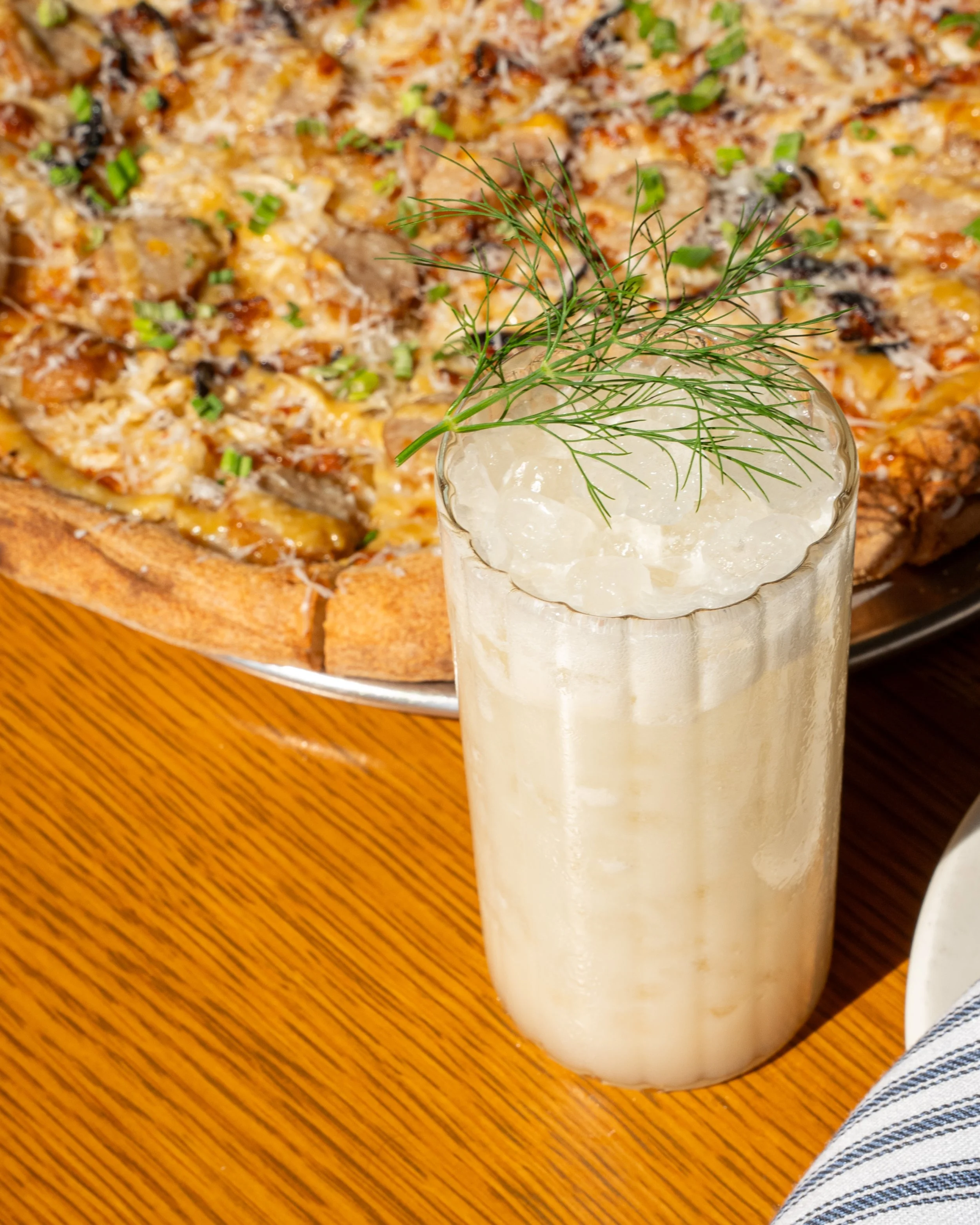 A glass of a creamy beverage with ice and a sprig of dill on top, next to a pizza on a wooden table.
