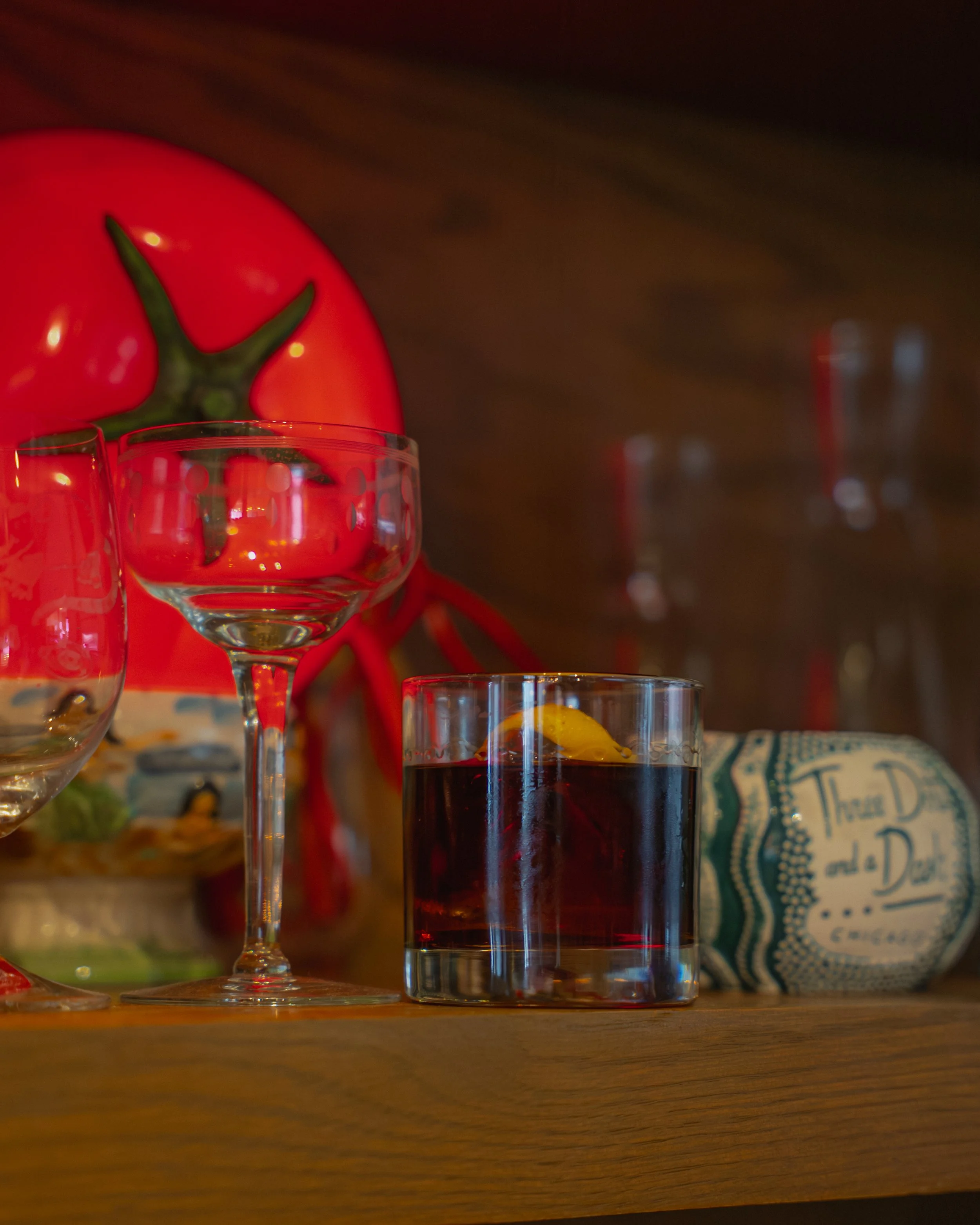 Close-up of a wooden shelf with a glass of dark red beverage with a lemon wedge, an empty wine glass, and a tomato behind them, along with a decorative object wrapped in paper.