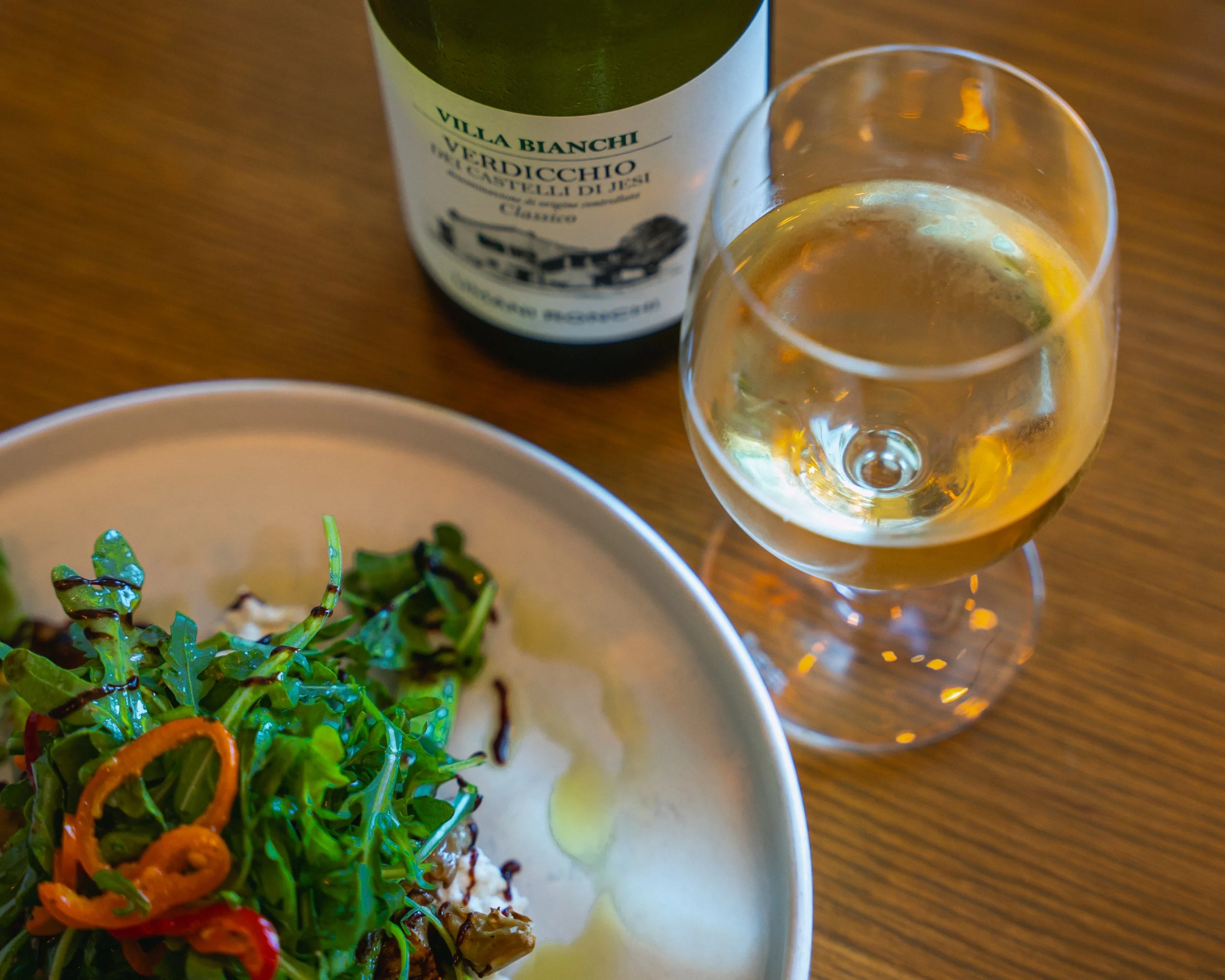 A glass of white wine, a bottle of Villa Banchi Verdicchio wine, a white plate with a mixed green salad topped with sliced red peppers, drizzled with balsamic vinegar, on a wooden table.