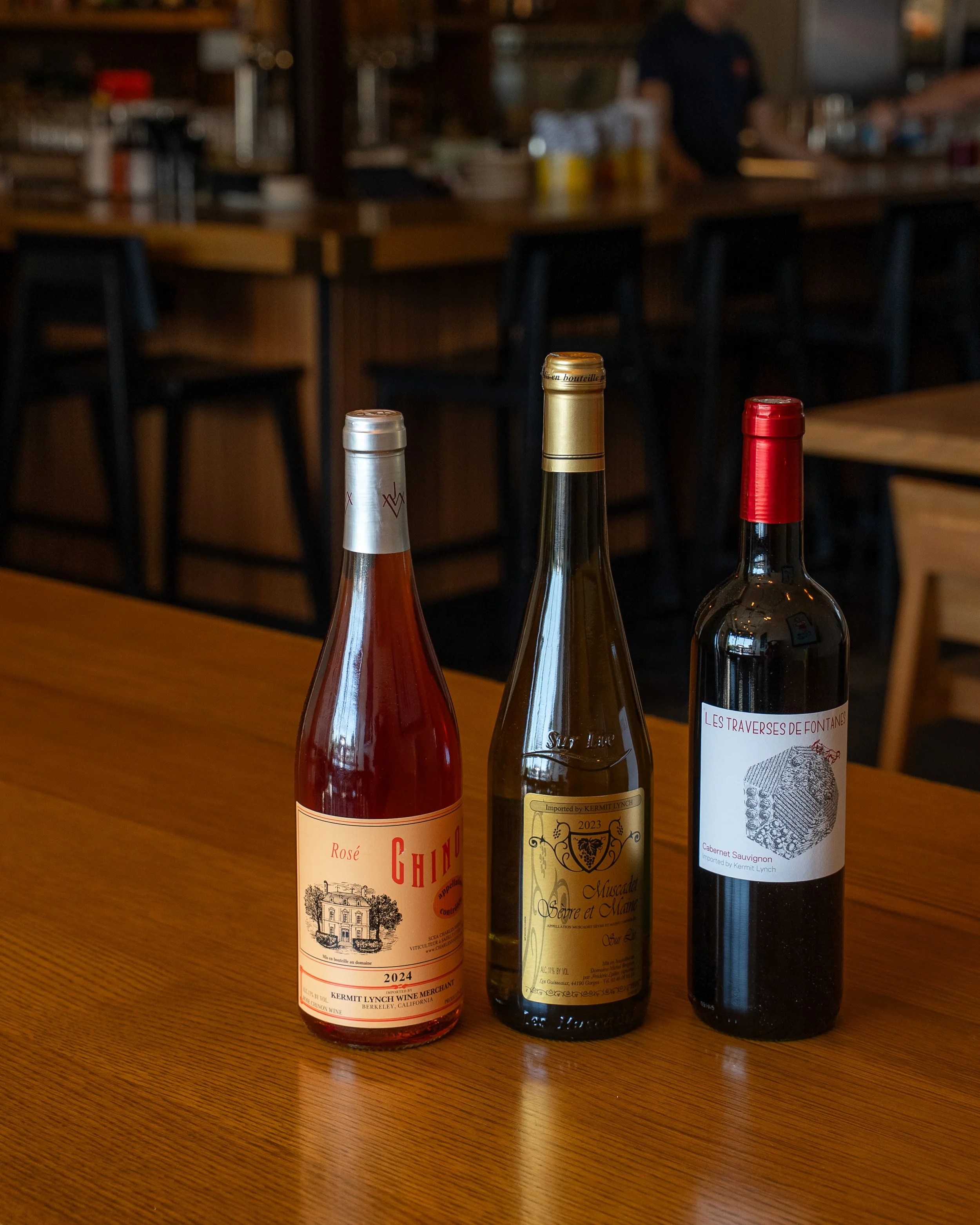 Three bottles of wine placed on a wooden table in a restaurant with a blurred background.